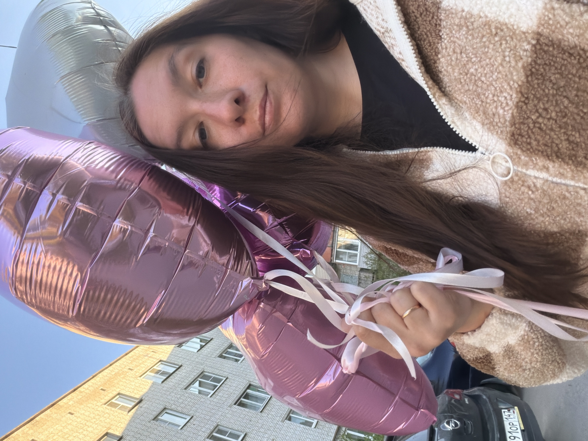 A woman with long brown hair is holding several large, metallic, heart-shaped balloons (primarily pink and one silver) with white ribbons tied to them. She is wearing a black shirt under a light brown, teddy-bear style jacket with a white zipper. She is holding the balloons in her right hand, which has a gold ring on the ring finger.
In the background, there is a multi-story building with multiple windows. The lower part of the building appears to be a pale yellow brick or stone, while the upper portion appears to be grey. A black car with a license plate visible (910P 142) is parked in the background. There is a hint of blue sky in the upper left corner. The general impression is of an outdoor urban environment, possibly Tokkari, Russia.