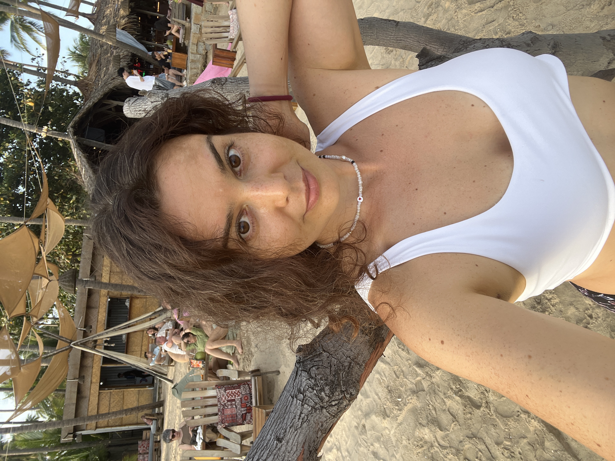 The image shows a woman taking a selfie on a beach. She is wearing a white bikini top and a beaded necklace with a small heart pendant. Her arm is resting behind her head, and she is leaning against a tree. In the background, there are several other people, beach chairs, umbrellas, and a small building that appears to be a bar or restaurant. The location is identified as Mũi Né, Vietnam. The beach has light-colored sand, and palm trees are visible in the background, suggesting a tropical environment. The sky is overcast, indicating it may be late afternoon or early evening.