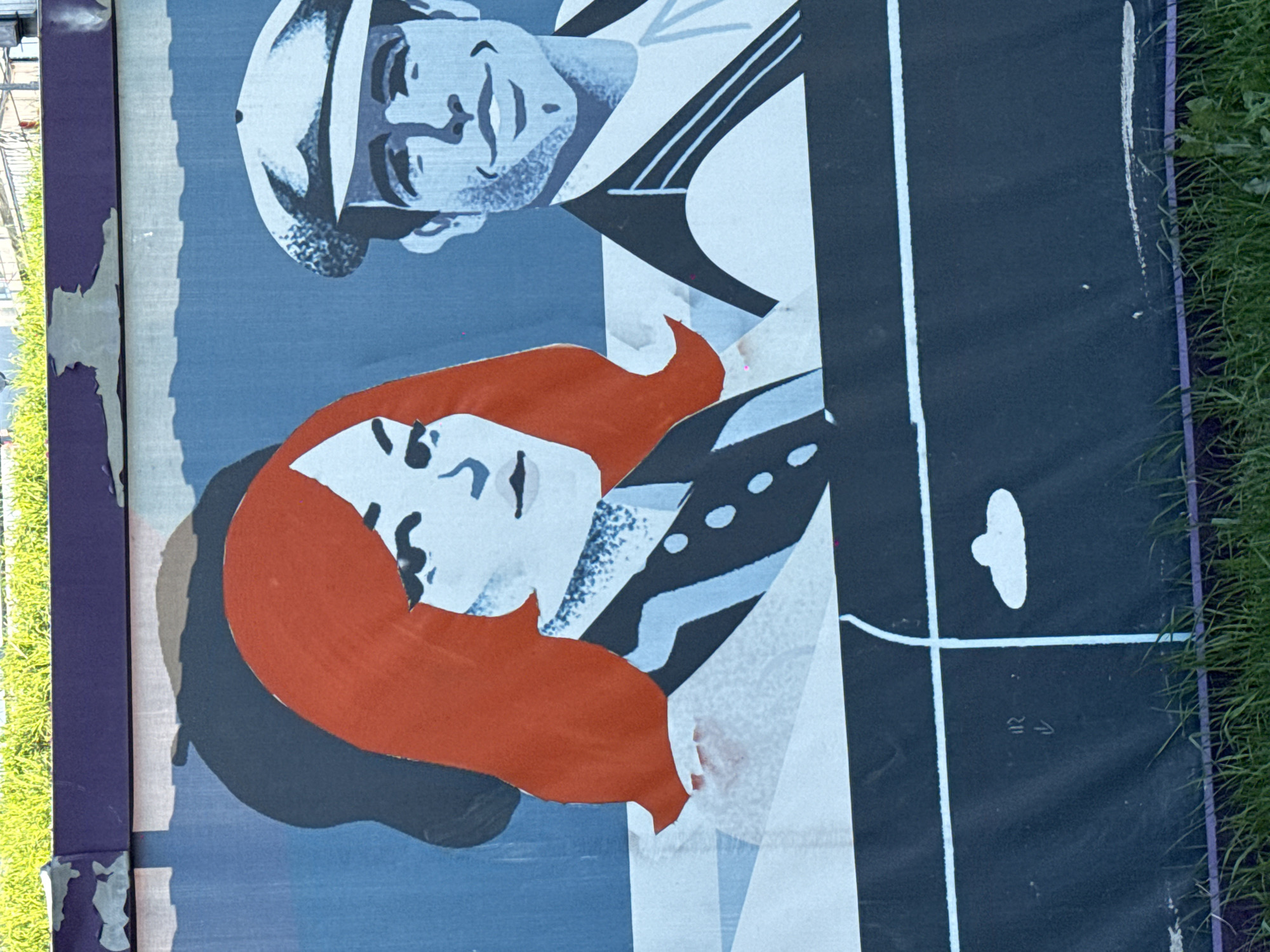 The image shows a portion of an outdoor advertisement or poster. It features a cartoon-like depiction of a man in a sailor's hat and a woman with red hair. The man is above the woman in the image. The background is a mix of blue, black, and white. There is grass visible on the right edge of the image. The poster appears to be damaged or peeling in places. The poster includes an arrow pointing to the upper left. The image was taken at an angle, suggesting it's a snapshot rather than a professionally staged photograph. The location is not clearly identifiable as Moscow, Russia, from this image alone.