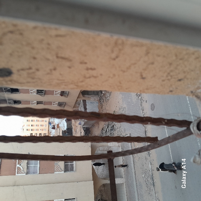 An elevated perspective captures a street scene. A metal railing runs across the foreground. Buildings with multiple windows line one side of the street. A street with a manhole cover is visible. People walk along the sidewalk. A white line is marked on the road. Debris and rubble are scattered along the edges of the street and around buildings. The image has a "Galaxy A14" watermark in the corner.