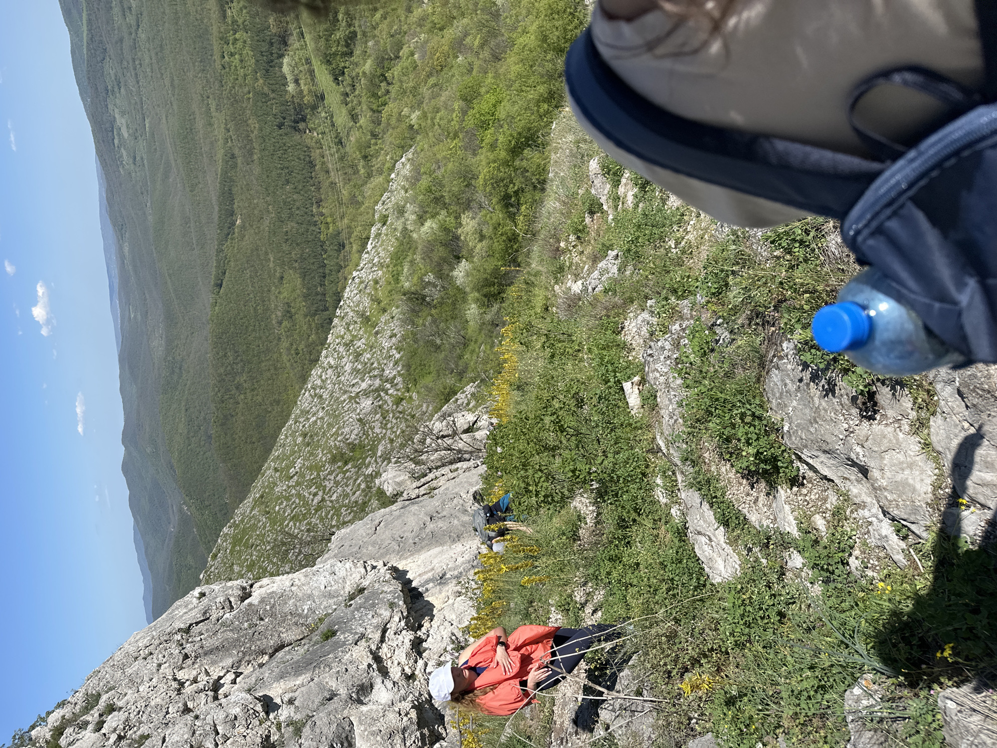 The scene is outdoors, on a rocky, grassy hillside in what appears to be a mountainous region, possibly near Bilka, Bulgaria, given the prompt's location. A woman lies on her back on the rocks and vegetation. She is wearing a white cap and an orange jacket. There is a backpack with a water bottle visible. The landscape slopes downward towards a distant, forested mountain range under a clear blue sky. There is another individual higher up on the slope. The perspective suggests a high vantage point or steep terrain.
