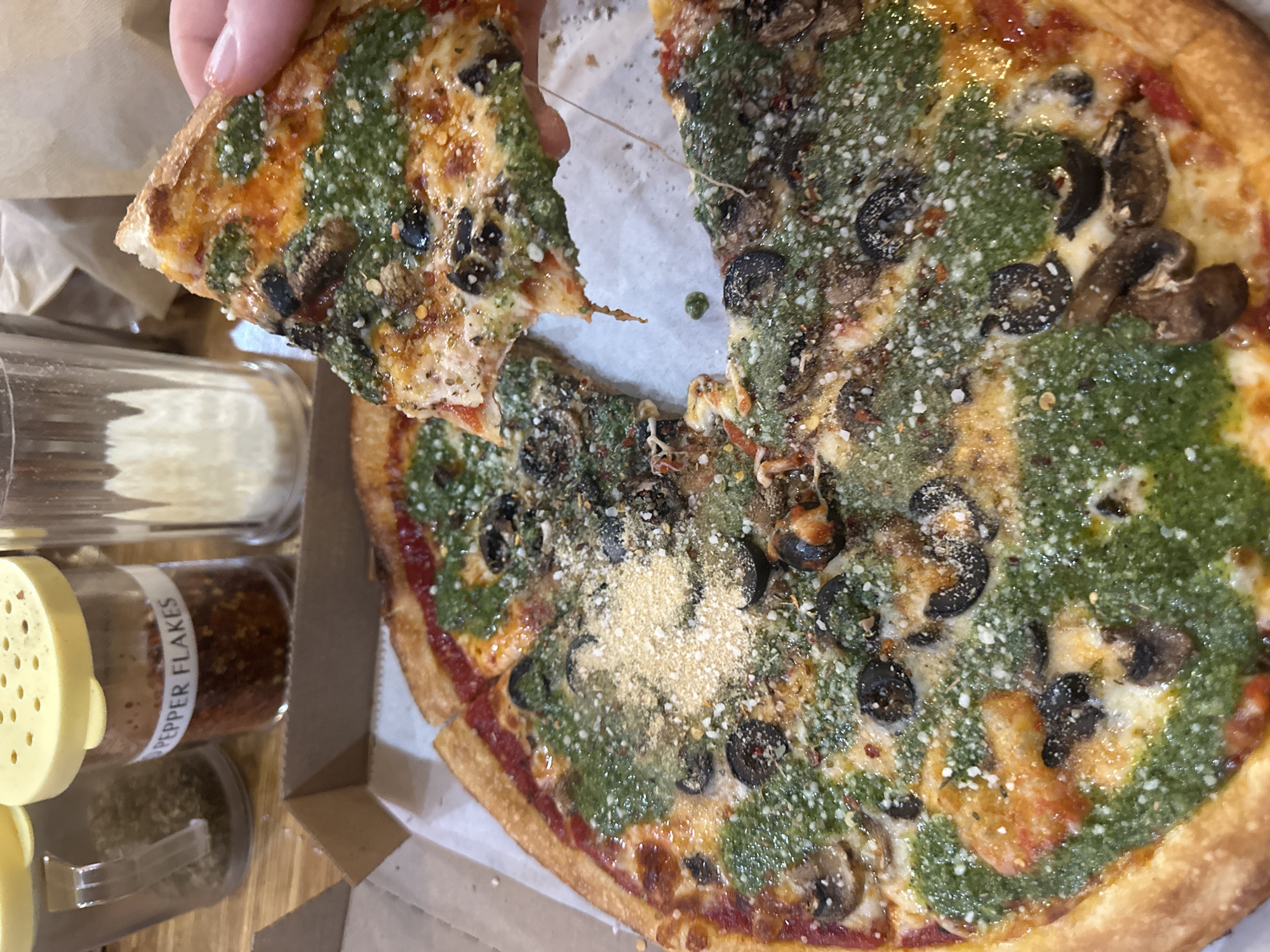 A close-up, high-angle shot captures a partially eaten pizza in what appears to be a casual dining setting in College Park, United States of America. A hand is seen in the upper left, lifting a slice of pizza, causing a string of melted cheese to stretch between the removed slice and the main pie.
The pizza itself features a vibrant green pesto sauce covering much of its surface, interspersed with patches of red tomato sauce and melted white cheese. Various toppings are visible, including sliced black olives and brown mushrooms scattered across the pizza. A generous dusting of grated Parmesan cheese and red pepper flakes adds texture and color to the top. The crust is golden brown and appears crispy.
The pizza rests in a cardboard box, with portions already missing, indicating it has been enjoyed. To the left of the pizza, several spice shakers are visible on a wooden surface. One clear glass shaker with a yellow lid, positioned vertically, clearly displays the text "PEPPER FLAKES" on its label, containing reddish-brown flakes. Another, larger shaker, possibly containing grated cheese, is partially visible behind it. The overall scene suggests an enjoyable meal, likely indoors.