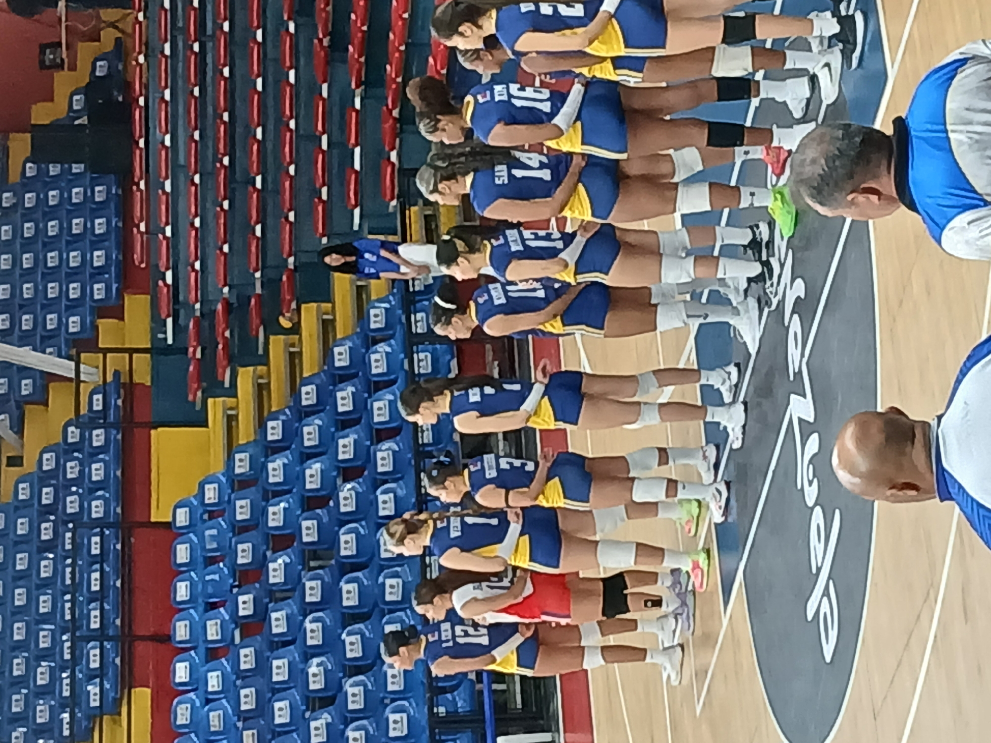 The image shows a volleyball team lined up on a court, likely for a game or practice. The players are wearing blue and yellow jerseys with numbers on their backs. There are rows of blue and red seats visible in the background, indicating that this is an indoor stadium. A man in a blue and white shirt stands on the court, presumably a coach or official. The letters "CXV" are visible on the court near the team. The location is Ciudad Guayana, Venezuela.