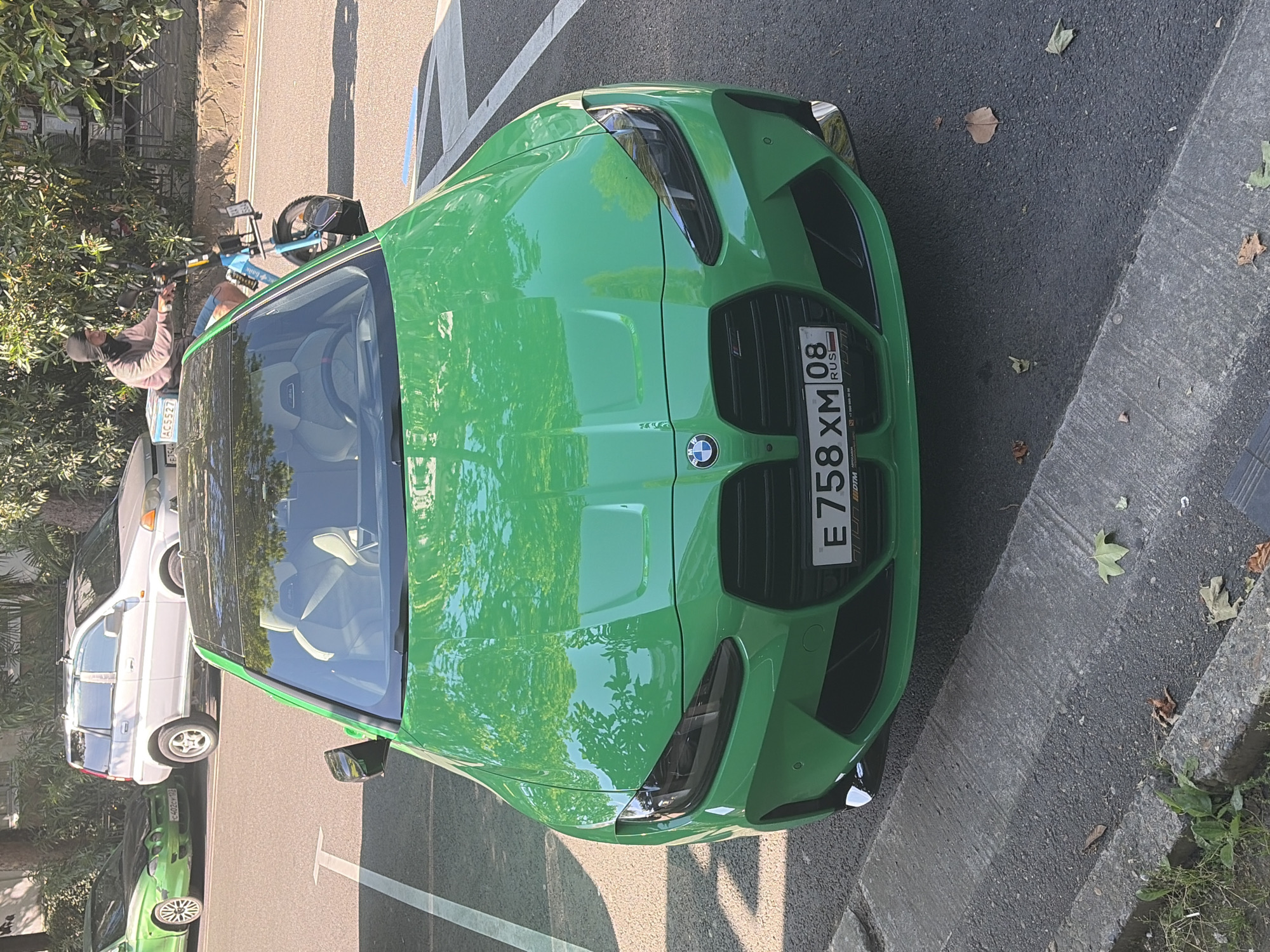 This image captures an urban street scene in Sochi, Russia, during daylight hours on a sunny day, characterized by strong shadows and reflections. The primary subject is a bright green BMW G82 M4 Competition parked on a paved surface, partially within a white-lined parking space. The vehicle features a black carbon fiber roof, black kidney grilles, dark headlights, and visible light-colored (likely white or light grey) carbon bucket seats through the windshield. Its Russian license plate reads "E 758 XM 08 RUS".
To the left and rear of the BMW, an older model green Hyundai Coupe (possibly a first-generation Tiburon) is parked, displaying a dark, potentially unpainted or damaged, hood and a Russian license plate "C 402 EY 93". Further behind the Hyundai, a silver first-generation Honda HR-V SUV is parked, equipped with a roof rack and partially visible Russian license plate "A 552 AC 93".
On the right side of the frame, a person wearing a black balaclava or head covering, a light brown long-sleeved shirt, and blue shorts is riding a light blue electric bike or scooter. This e-bike has thick tires and a white delivery box affixed to its rear rack. The rider is looking towards the left side of the frame.
The environment consists of an asphalt street with white painted lines. A concrete curb runs along the immediate foreground and to the right of the green BMW, separating the road from a strip of ground with sparse grass, weeds, and scattered brown and green fallen leaves. Lush green trees and bushes form a dense backdrop, reflecting prominently on the hood of the green BMW.