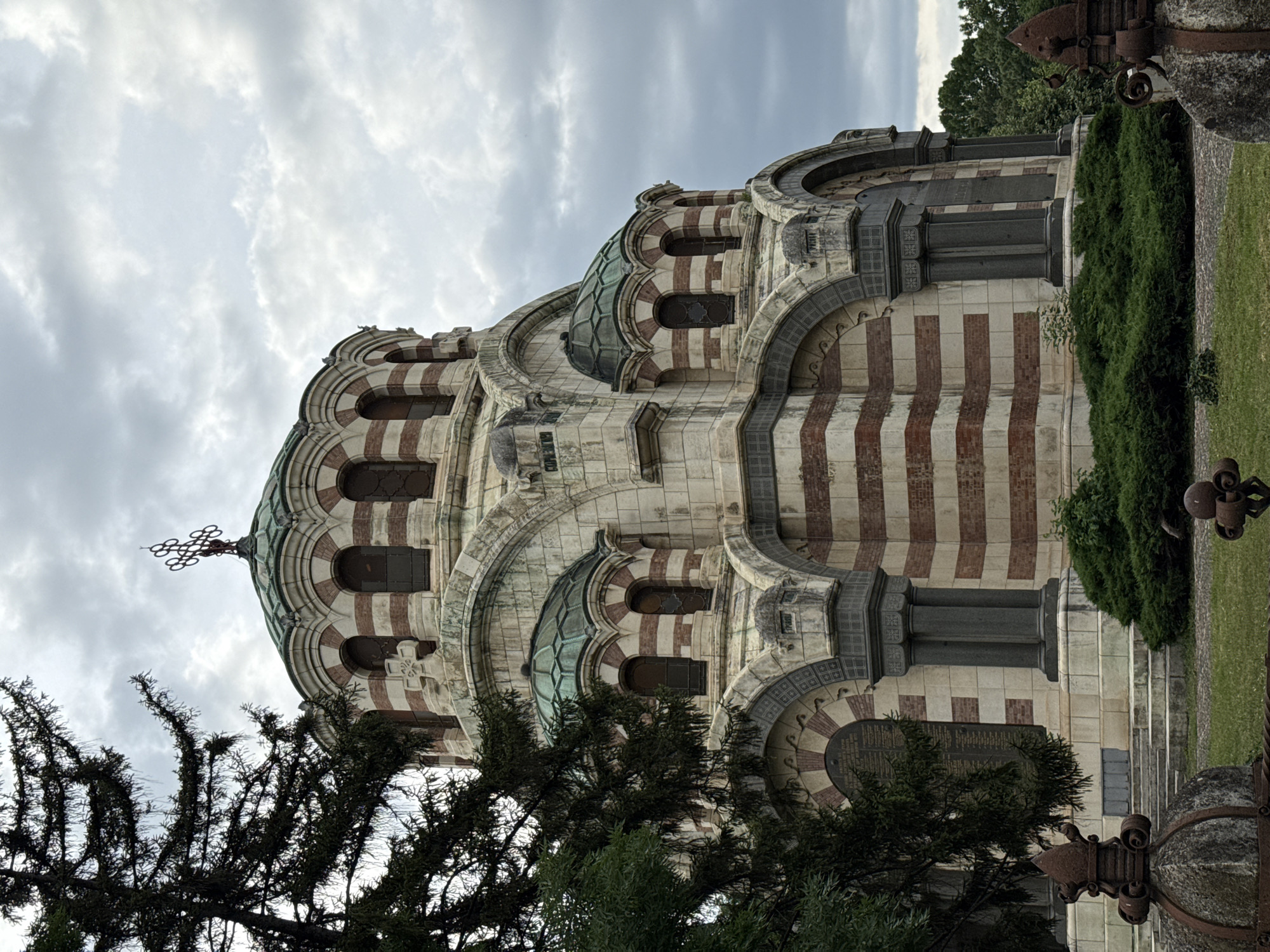 The image shows a view of the Pleven Mausoleum, also known as the St. George the Conqueror Chapel Mausoleum, located in Pleven, Bulgaria. The mausoleum is a domed, octagonal structure constructed with alternating bands of light-colored stone and red brick. It features multiple smaller domes and a prominent large dome topped with a cross. The architecture exhibits Byzantine influences. In the foreground, to the left, a large evergreen tree partially obscures the view of the mausoleum. The grounds around the mausoleum are grassy, with some low-lying green bushes in front of the structure. In the lower portion of the image, there are decorative iron fence elements, some of which appear rusted. The sky is overcast with heavy gray clouds. There are no people visible in the image.