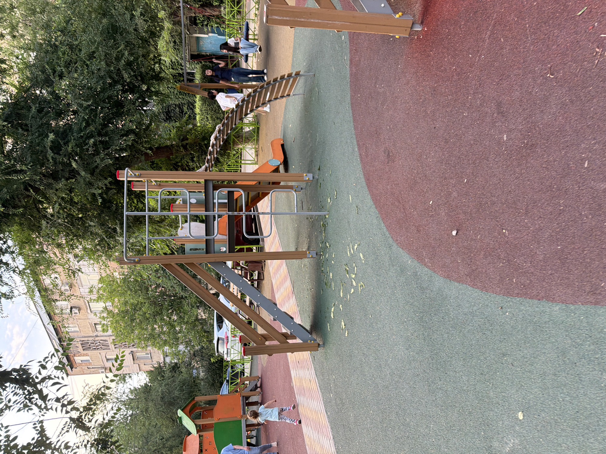 The image shows a public playground in Shymkent, Kazakhstan, likely in a residential area given the apartment building in the background. The playground features a modern play structure with a wooden staircase, a rope bridge, and an orange slide. A young child in striped leggings is running on the rubberized surface of the playground. In the background, three young women are standing near a swing set, with two of them interacting and one looking away. The environment is partially shaded by large trees, and the ground surface is a mix of green and red rubber tiles. The apartment building behind the playground appears to be an older Soviet-era structure, common in many cities in Kazakhstan. The overall scene depicts a typical urban playground scene in the late afternoon or early evening.
