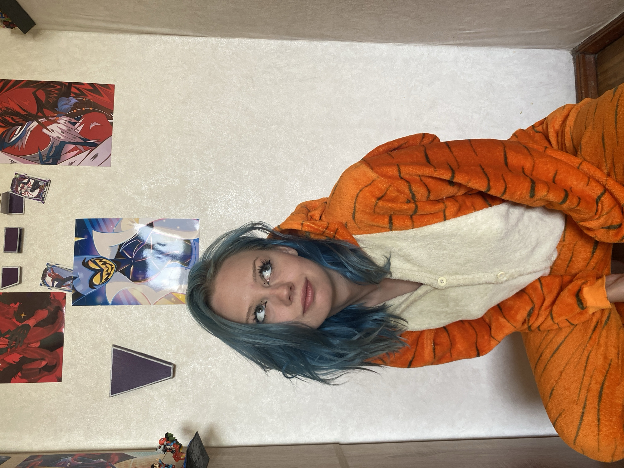 A young Caucasian woman with short, blue hair is sitting indoors, wearing an orange and white tiger-striped onesie. Her eyes are looking upward and to the right, and she has on eyeliner. She is positioned in front of a white, textured wall. Several framed pictures and small figurines are displayed on the wall behind her. To the left, a sliver of a wooden closet door is visible. The room appears to be a personal space, possibly a bedroom or living area, based on the decorative elements and furniture. The lighting is soft and natural, coming from an unseen source.