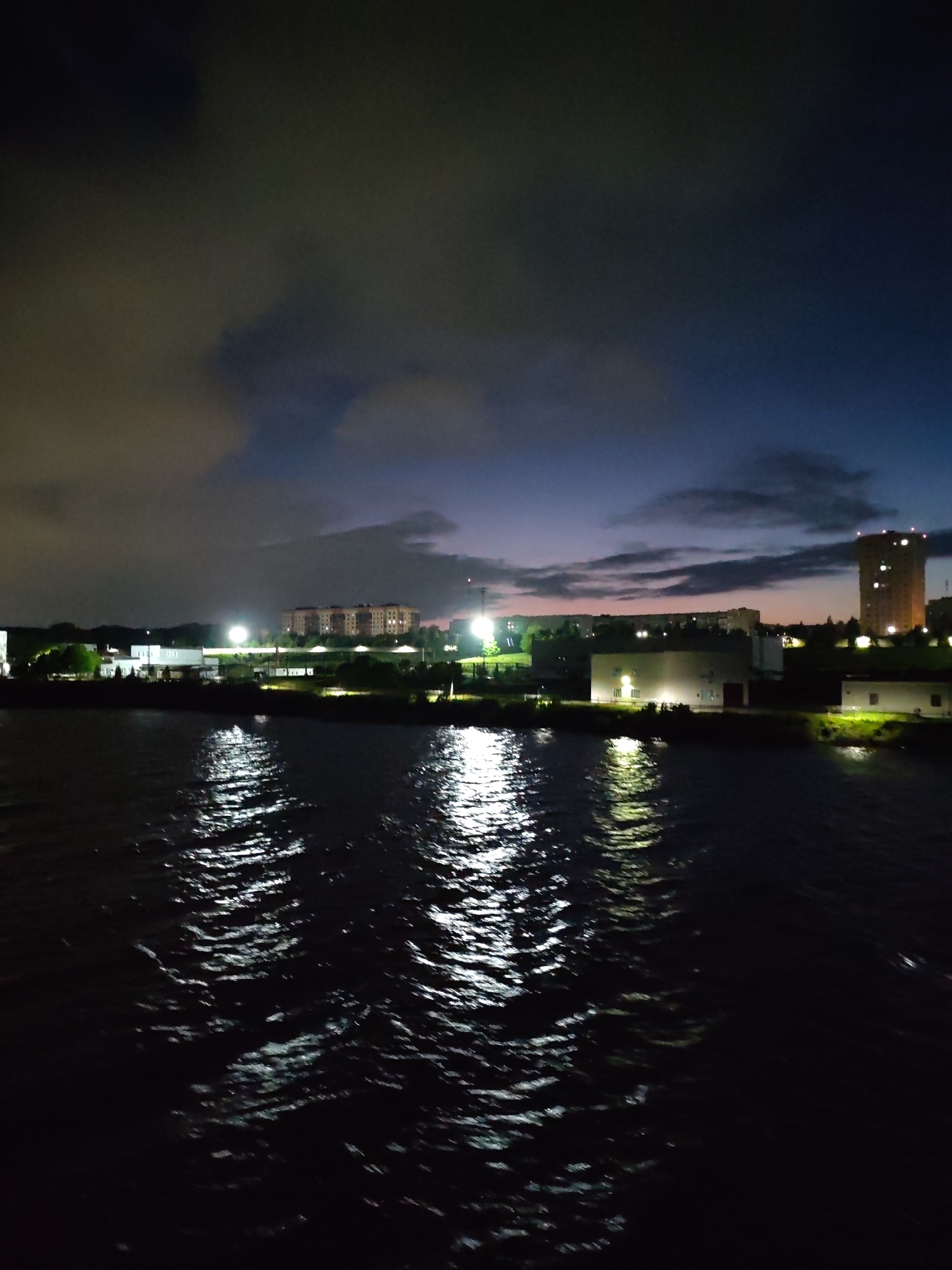 At night, the cityscape of Cherepovets, Russia, is seen across a body of water, likely the Sheksna River. Reflected city lights create shimmering patterns on the water's surface. Buildings illuminated at night stand along the horizon, including what appears to be residential complexes and industrial structures. A tower-like building with lights is visible on the right side of the image. The sky is a mix of dark blue and shades of grey from the clouds.