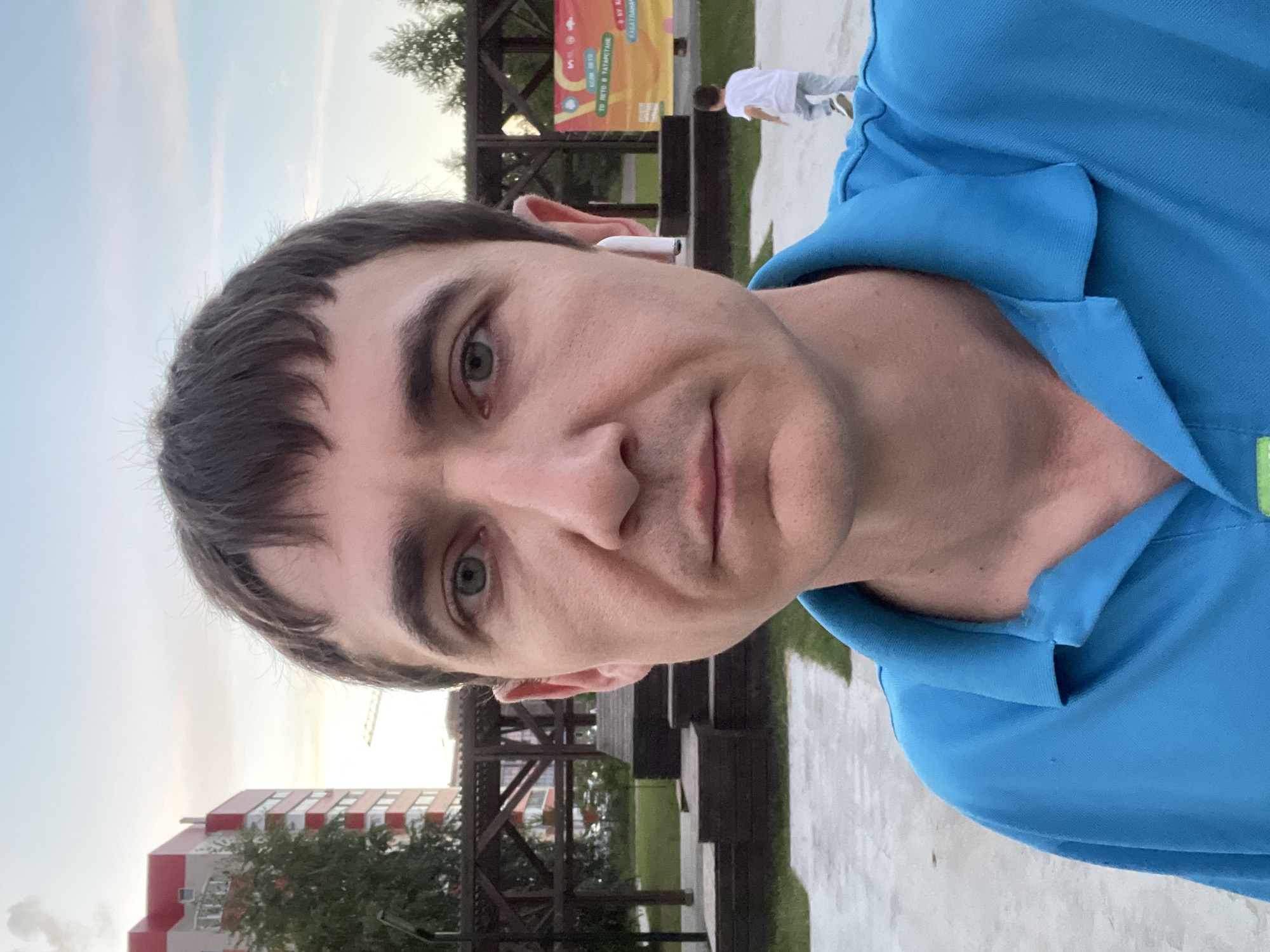 A man with short, dark hair and blue eyes takes a selfie outdoors. He is wearing a blue polo shirt and a white wireless earbud in his right ear. The background shows a park-like setting with a wooden playground structure, green grass, and a building with red accents. A young child in a white t-shirt and grey pants is running in the background. A banner with text and colorful graphics is visible on a fence behind the child. The sky is overcast.