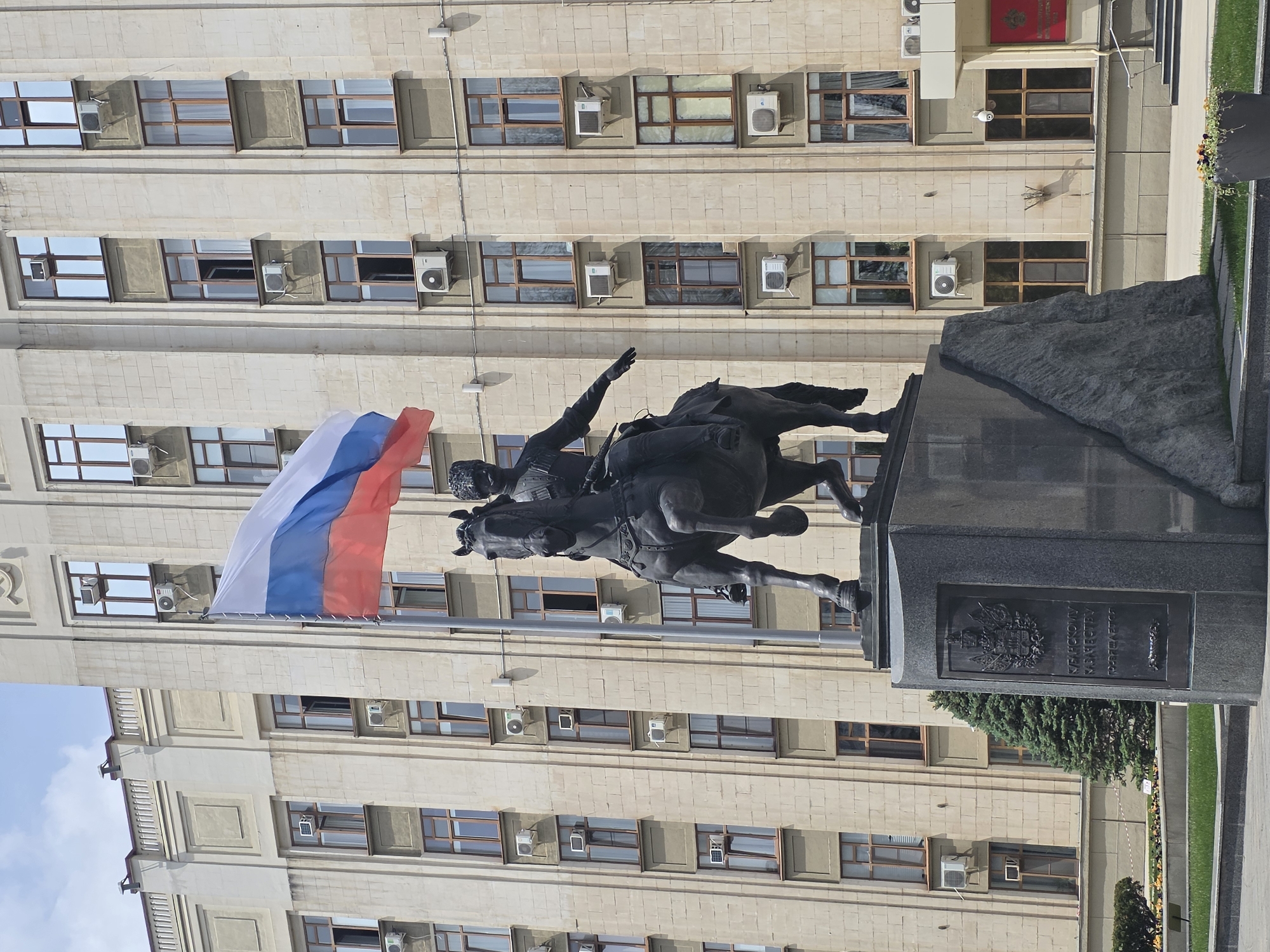 The image depicts a statue of a Cossack on horseback in front of a building in Krasnodar, Russia. The statue is dark and appears to be made of metal. The Russian flag is visible near the statue. The building behind the statue is a large, multi-story structure with many windows. The sky is visible in the top left corner of the image. There is also grass and flowers to the side of the statue.