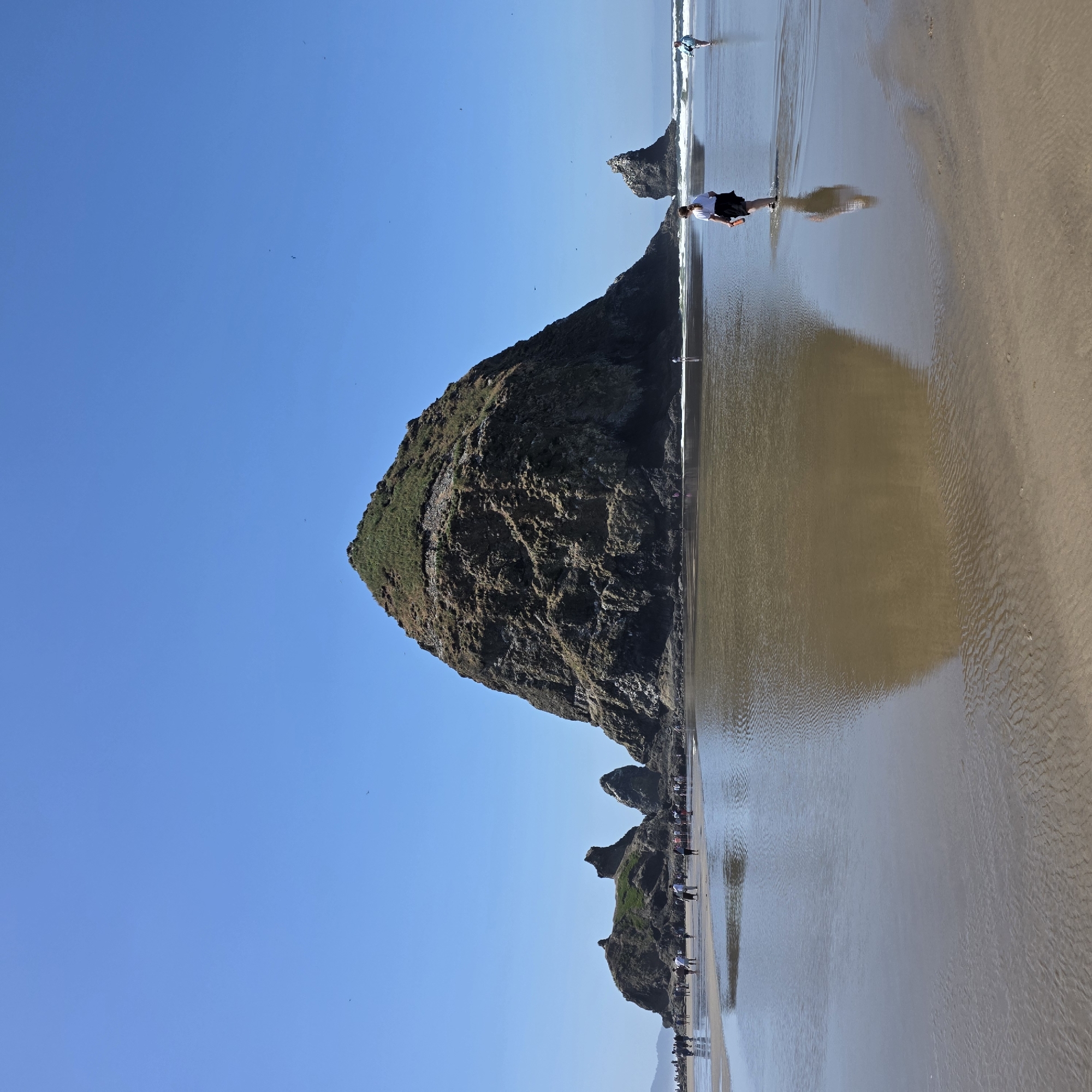 This is a landscape photograph taken at Haystack Rock in Cannon Beach, Oregon, United States of America. The image depicts a wide, flat beach with shallow pools of water reflecting the sky and the iconic rock formation. In the foreground, a young girl with blonde hair tied in a ponytail, wearing a white t-shirt and a dark skirt, is walking through the water, holding an orange object. Her reflection is clearly visible on the wet sand. Further into the scene, other people are scattered across the beach, some walking, others standing near the water's edge. To the left, smaller rock formations are visible, with a larger, prominent haystack-shaped rock dominating the center of the frame. This rock is covered in greenery and appears weathered. The sky is a clear, bright blue with a few faint specks that could be birds. The overall atmosphere is serene and suggests a pleasant day at the beach.