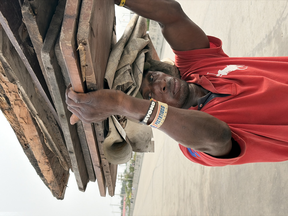 A man in a red Ralph Lauren polo carries a stack of weathered wooden planks on his head. The load is balanced with the help of a beige cloth underneath. He wears two wristbands on his right arm, one reading "BEYOND," the other reading "EXPECTATIONS." He gazes directly at the camera with a weary expression. The background shows a hazy cityscape.
