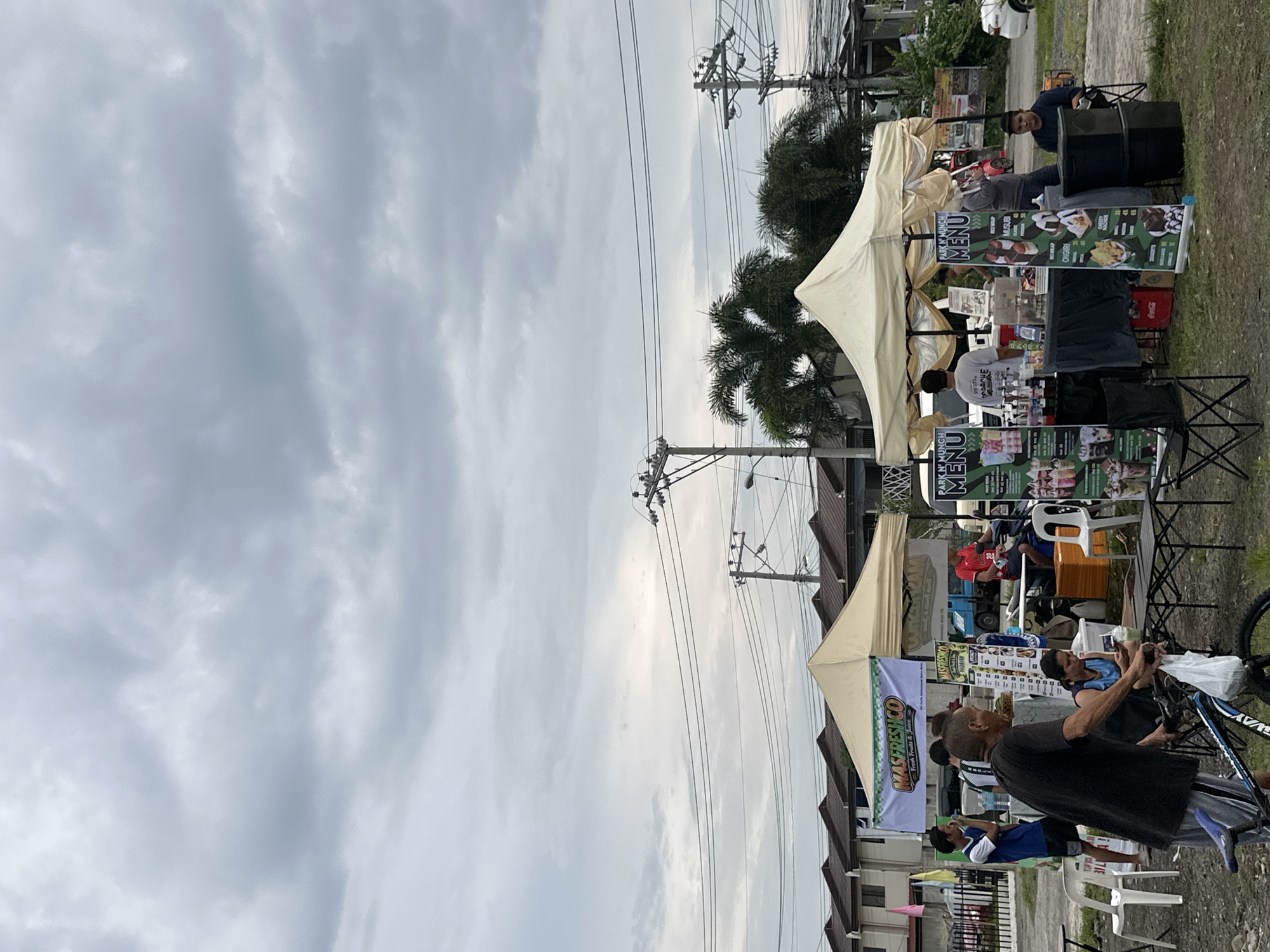 The image depicts an outdoor food market or street fair in Davao City, Philippines, under an overcast sky. The scene is bustling with several pop-up stalls and people.
In the foreground, a man in a black t-shirt is seen from behind, holding a bicycle, standing near a food stall. This stall, on the left, features a green banner for "MASFRESHCO Fresh Fruits & Juice." The central and right portions of the image showcase multiple food stalls, each covered by a light-colored canopy. Several "PARK N' MUNCH MENU" banners are prominently displayed, listing food items such as "ONGIRI" and "MUSUBI" with prices like "45" and "65," indicative of local Asian-inspired cuisine.
Behind one of the stalls, a man in a dark blue shirt and cap smiles towards the camera, positioned next to a black drum. Another individual in a white t-shirt attends to customers at an adjacent stall. Folding chairs are scattered around, with some occupied by patrons.
The background features numerous utility poles with crisscrossing electrical wires, characteristic of the region. Palm trees are interspersed among the stalls and residential buildings that line the area. The ground appears to be a mix of grass and dirt, with a white car partially visible in the distance. The cloudy sky suggests either an overcast day, late afternoon, or early evening. The overall atmosphere is casual and community-oriented, reflecting a local gathering or a regular outdoor market setup.