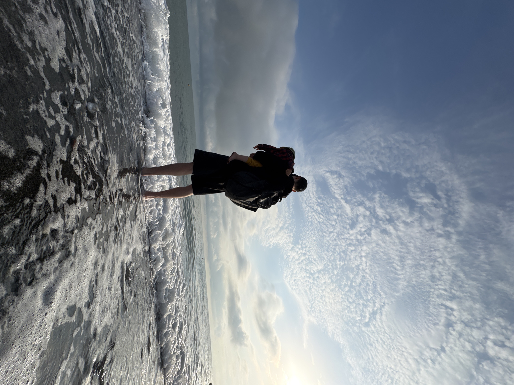 A father holds a child on a beach in Sochi, Russia. The father stands in the shallow surf, wearing black shorts and a black jacket. The child, wearing a red and black plaid shirt and yellow pants, is held facing away from the camera. The sky is partly cloudy, with a mix of blue sky and grey clouds. The beach is dark sand, and the water is foamy with waves. The sun shines in the lower part of the image.