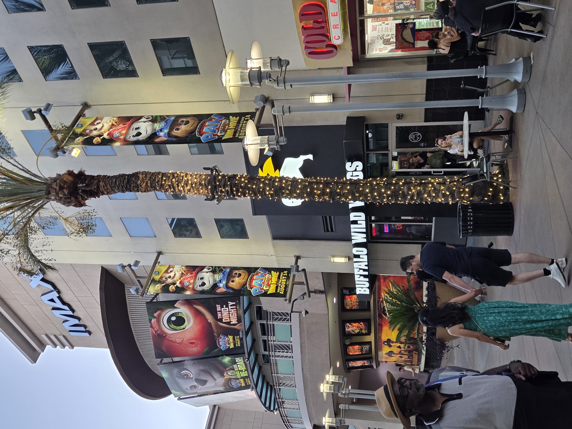 The image shows a street scene in Burbank, California, in front of an IMAX theater and a Buffalo Wild Wings restaurant. The IMAX theater features large banners advertising the "PAW Patrol: The Dino Movie". A man in a straw hat and glasses is in the foreground, looking to the left. A couple, a man and woman holding hands, are walking away from the camera. Several people are seated at tables outside Buffalo Wild Wings, and a woman is sitting alone at a table, looking at her phone. A palm tree decorated with string lights is prominent in the image. The sky is overcast.