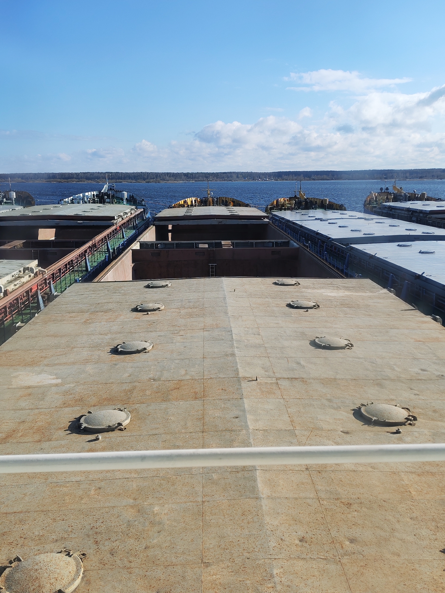 The image shows a view from the deck of a barge or ship in Bol'shie Mkhi, Russia. The foreground is dominated by a rusty, flat, concrete deck with circular access hatches. A white railing is visible along the lower edge of the frame. The barge is seemingly docked alongside at least three other similar vessels, with visible walkways between them. The background features a body of water, possibly a river or lake, with a treeline along the distant shore under a partly cloudy blue sky.