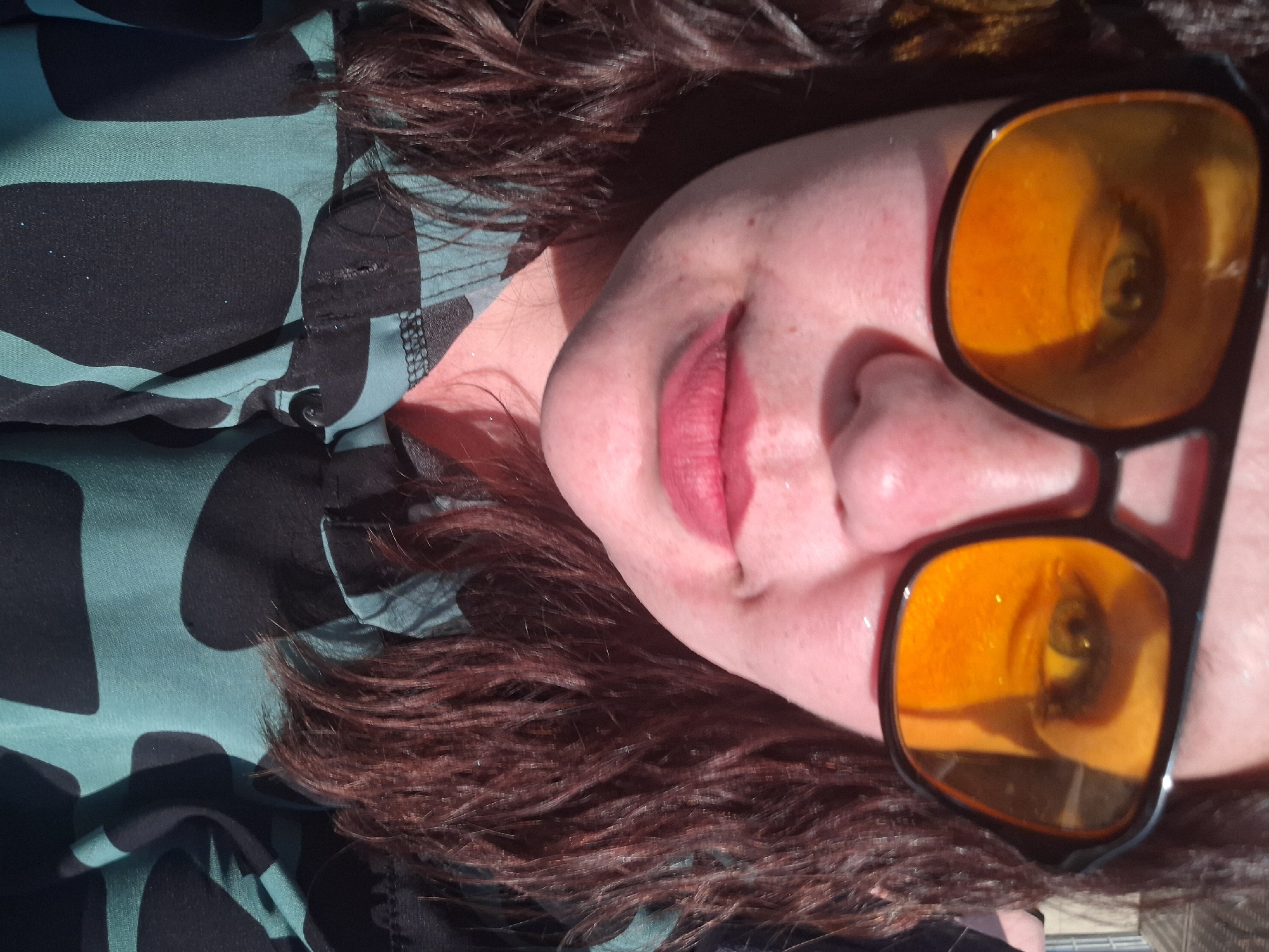 A close-up photograph depicts a person lying down with their head turned to the side. The person has fair skin, pink lips, and brown curly hair. They are wearing sunglasses with orange lenses and black frames. A green and black patterned shirt is visible on the left side of the image. The background is out of focus. The location is not discernible from the image, so it is not possible to determine if it is in Kamennyi Kliuch, Russia.