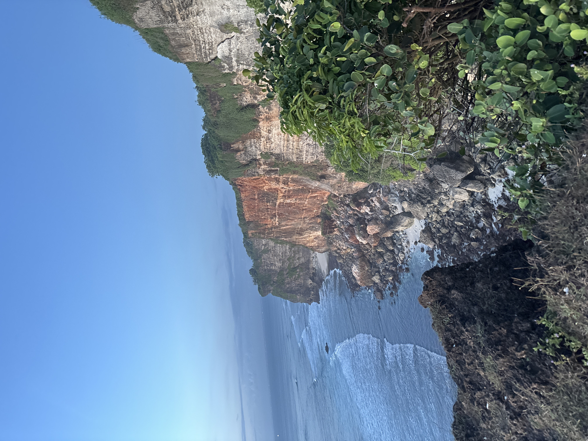 The image depicts a coastal scene in Kuta Selatan, Indonesia, featuring a dramatic cliff overlooking the ocean. The cliff is composed of layered rock formations, exhibiting varying colors and textures, with vegetation growing along its top edge. Below the cliff, the ocean waves crash against the rocky shore. The sky is clear and blue.