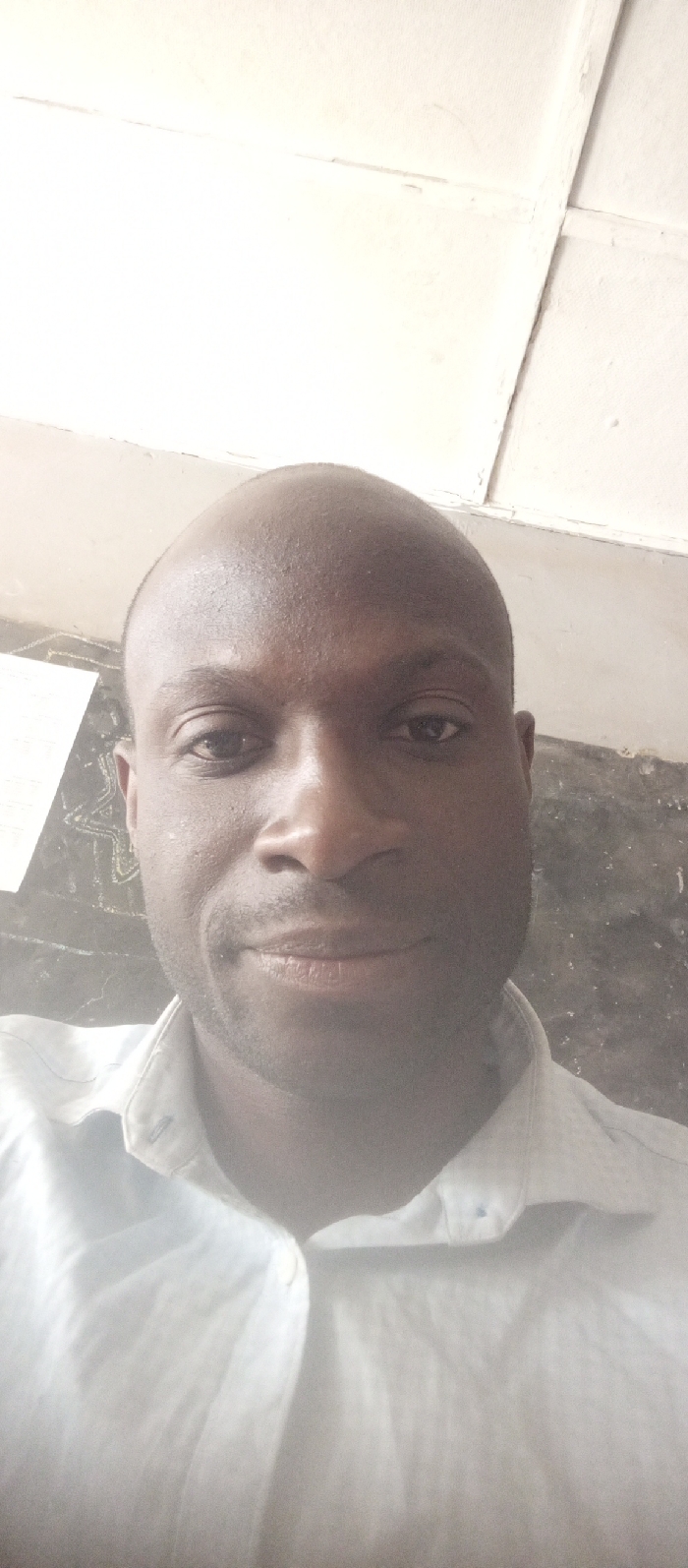 A close-up, head-and-shoulders portrait of an adult male is captured indoors. The subject, of African descent, has a bald or closely shaven head and a visible stubble on his chin and cheeks. He wears a light-colored, possibly white or light blue, collared button-up shirt with the top button unfastened. His eyes are open, and his mouth is closed, forming a slight, upward curve, indicative of a subtle smile. He is looking directly forward, into the camera. The background to the subject's left (viewer's right) reveals a dark, textured wall with some indistinct, lighter markings. Above the subject and to his right (viewer's left), a light-colored, possibly white, panelled ceiling with visible seams and some discoloration is present. The lighting is bright, causing some overexposure on the upper portion of the subject's head and the ceiling. The scene is located in Barnawa, Nigeria.