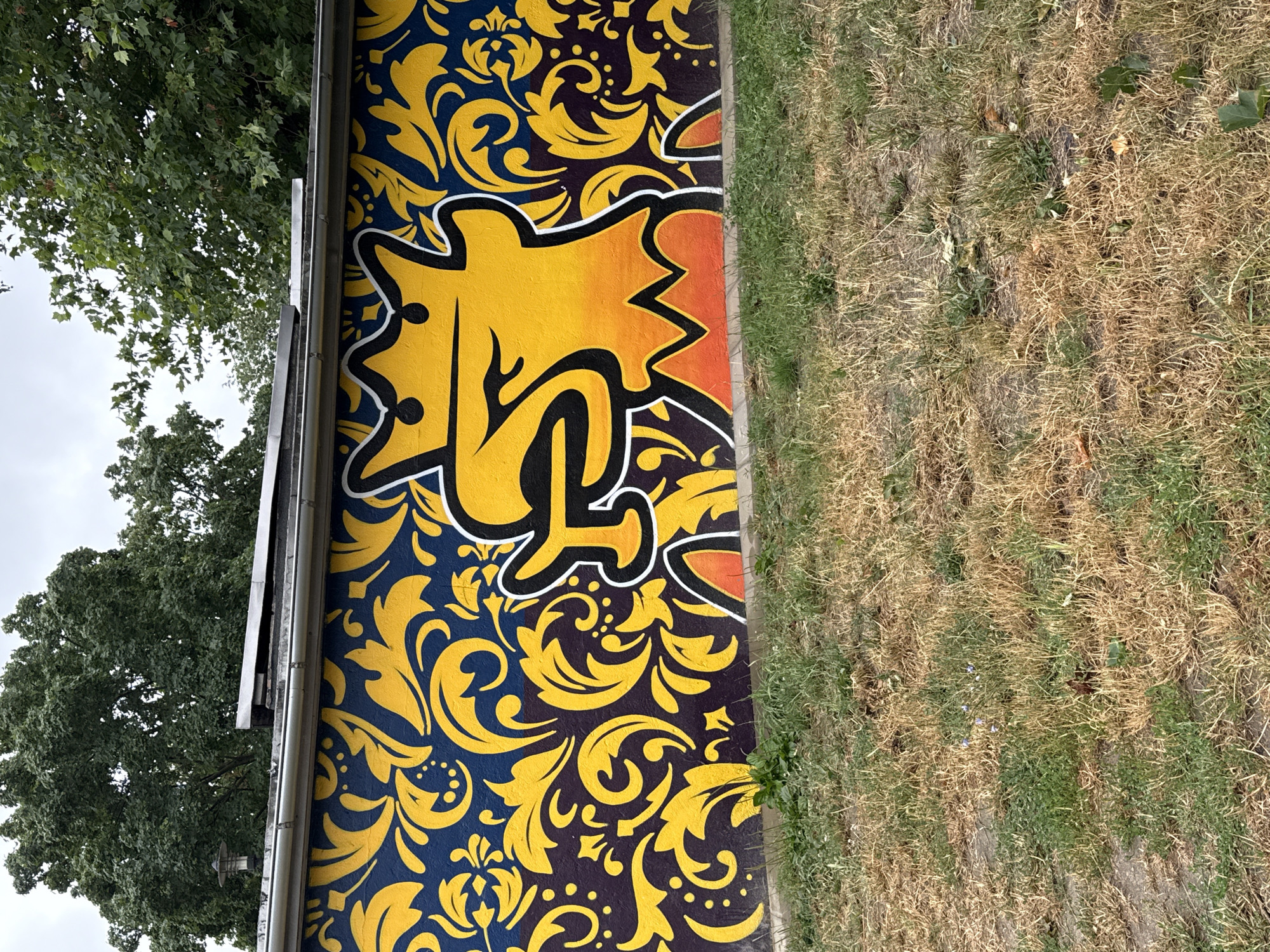 This media file shows a section of a mural painted on an exterior wall. The mural features a vibrant design with a central yellow graphic element outlined in black and white, set against a dark blue background filled with intricate yellow, ornate floral patterns. The yellow graphic appears to be a stylized, abstract representation of a figure or creature, possibly with a crown-like shape at the top. The mural is bordered on the right by a strip of dry, light brown grass, indicating an outdoor setting in what might be a park or a neglected urban area. To the left of the mural, a portion of a mature tree with lush green foliage is visible, suggesting a natural element within the environment. A metal gutter runs along the top of the wall where the mural is painted, hinting that this could be the side of a building. The sky appears overcast, suggesting a cloudy day. There is no visible text in the image. The overall impression is one of urban art in an outdoor environment, contrasting the colorful, stylized artwork with the natural and somewhat unkempt surroundings. The location context provided is Szczecin, Poland.