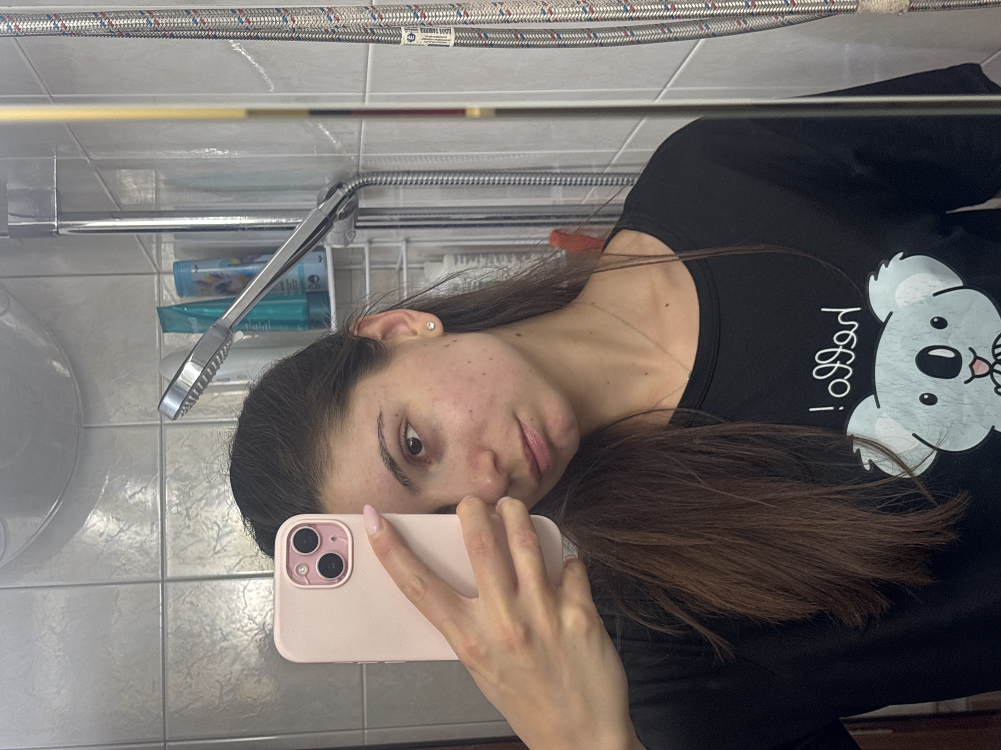 The image depicts a young woman taking a selfie in a bathroom. She is holding a pink smartphone in her right hand, capturing her reflection in a mirror. The bathroom appears to have tiled walls and a shower fixture is visible in the background, along with bottles of toiletries on a shelf. The woman has long, dark brown hair and is wearing a black t-shirt with a graphic of a koala. Her facial features suggest she is looking directly at the camera. The environment is brightly lit, likely from overhead bathroom lighting. There is no indication of the specific location within Varna, Bulgaria.