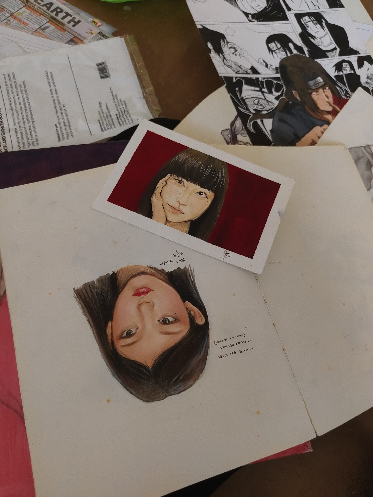 An overhead, slightly angled shot displays an art workspace featuring various papers and drawings. Centrally, a large open sketchbook page lies flat, showcasing an upside-down, colored portrait drawing of a young woman with dark hair and red lips. Handwritten notes next to this drawing read "UNEVEN EYES," "MORE DETAILS," and "(LABI NA SA EYES)" along with "ELI 12/18/23." Overlapping the top edge of the sketchbook is a smaller, finished watercolor or gouache painting on a white card, depicting another young woman with dark hair and bangs, resting her chin on her hand, against a solid red background. A faint signature is visible below the woman's face on the small painting.
To the upper left, a document or publication titled "EARTH" and "AND DISCOVER" is partially visible. Beneath this, a white plastic-wrapped package label for "BATH AND SHOWER GLOVES" is present, displaying product information and a barcode. The label explicitly states "Distributed by: Soffiya Skin Care Products... Davao City, Philippines" and "Manufactured for: Soffiya Skin Care Products, Inc... Davao City 8000, Philippines." To the upper right, a sheet of paper or comic book page is folded, displaying multiple black-and-white and one colored illustration of manga characters, notably Itachi Uchiha from Naruto. The overall surface appears to be a light-colored table or desk, showing minor smudges.