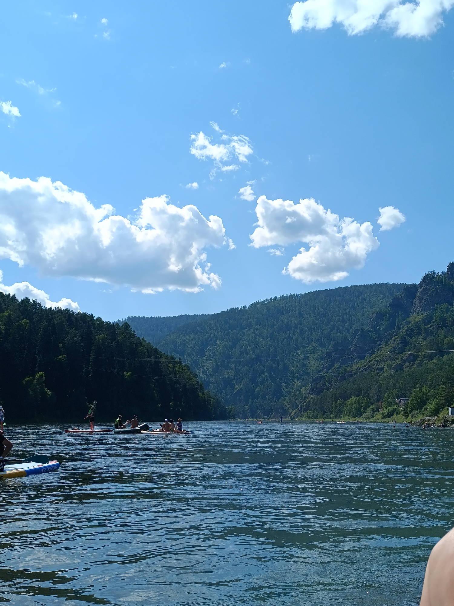 The image depicts a wide, blue river with rippling water under a clear sky with scattered white clouds. On the water, several people are engaged in stand-up paddleboarding. In the foreground, one paddleboard is visible with a person's leg on it. Further back, other paddleboards with multiple occupants are spread across the river. The riverbanks are lined with dense, green, coniferous forests that ascend into rolling hills and mountains. The scene is set in the city of Ust-Mana, Russia, under bright daylight.