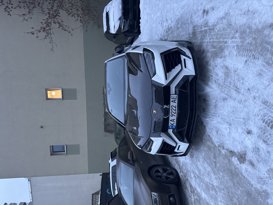The image shows a snowy street with cars parked alongside a building. A white Lamborghini Urus with a black hood and "KA 9222 AE" license plate is prominently featured. Another car is parked behind it, partially covered in snow. A brown sedan is visible in front of the Urus. The building has two windows; one has a warm, lit glow. Tire tracks mark the snowy ground. The scene gives a wintery urban feel.