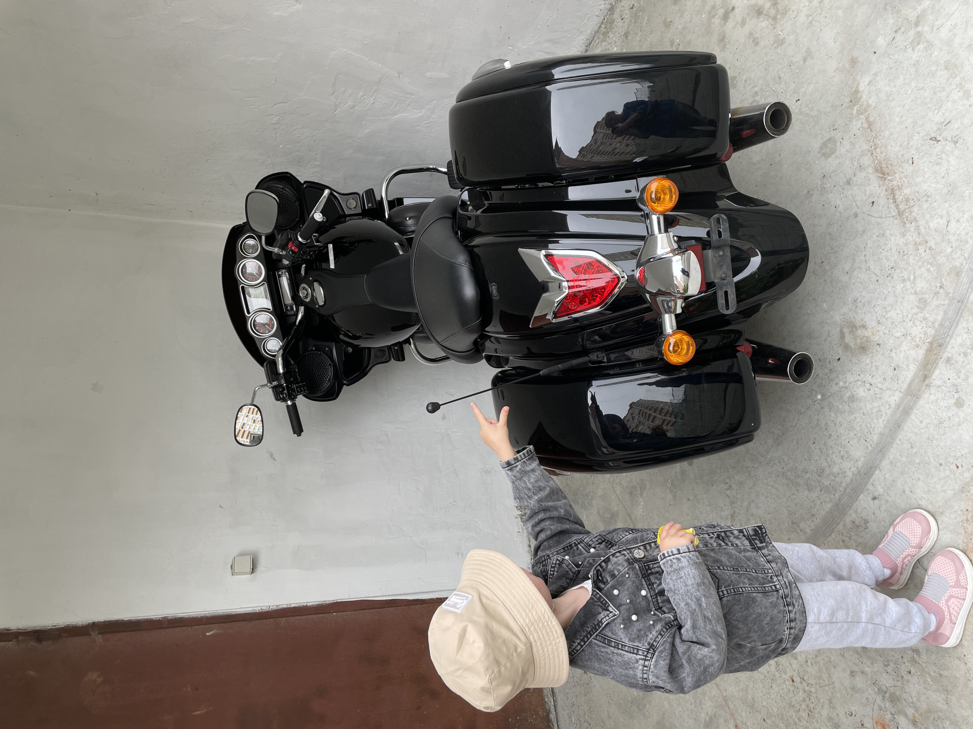 A child stands near a black motorcycle. The child wears a tan bucket hat, a gray denim jacket, gray sweatpants, and pink sneakers. The motorcycle has a large back storage compartment. The scene appears to be indoors, possibly a garage, with gray concrete walls and floor. A brown door is visible in the lower left corner. The photograph provides no information to suggest the city is Vladivostok, Russia.