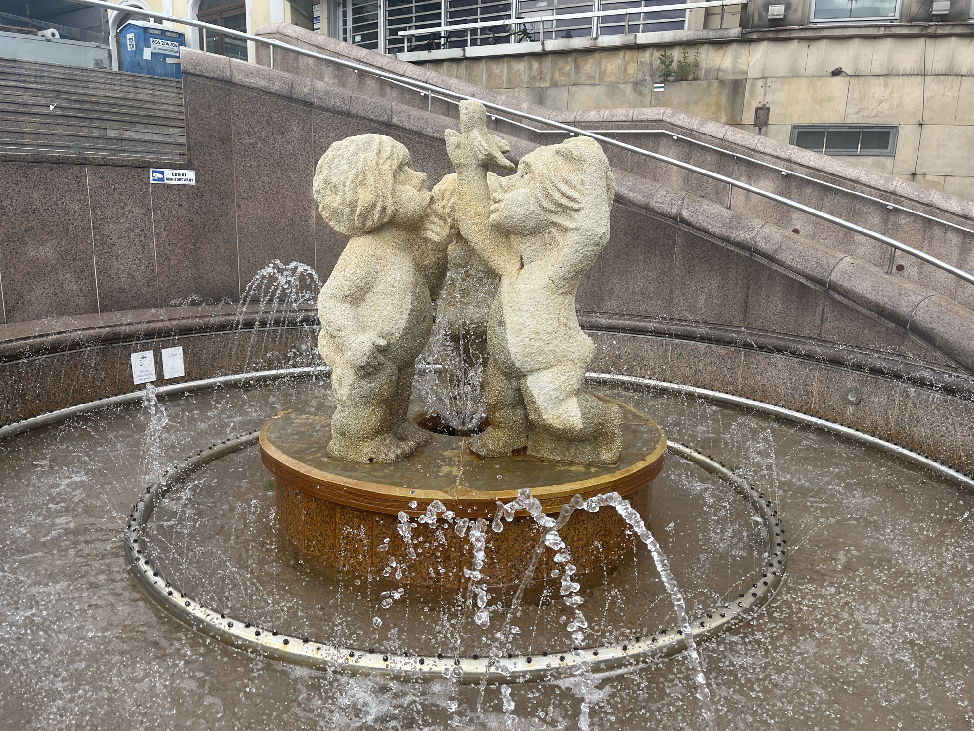 A stone fountain, located in front of a multi-story building with stone facade and a metal staircase, features two stone figures of children interacting with a small object. The figures are positioned on a circular wooden base within a pool of water. Water sprays from the pool's edge, creating a circular ripple effect. The environment appears to be a public outdoor space.