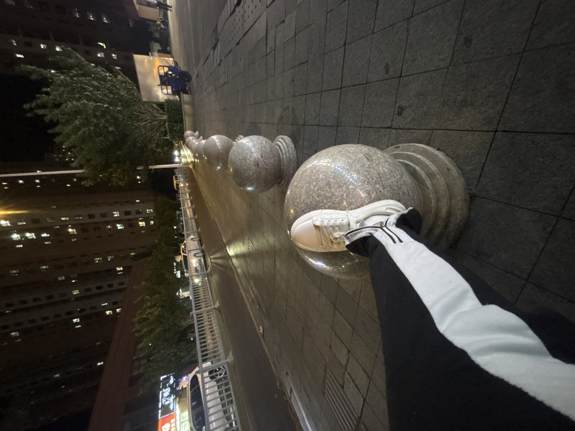 A person's left leg, wearing a black pant with a white stripe and a white sneaker, is resting on a rounded stone bollard on a paved sidewalk at night. In the background, a street is visible with cars and a white fence. Beyond the street, tall apartment buildings with illuminated windows are present. Trees line the area between the street and the buildings. A blue three-wheeled vehicle is parked on the right side of the sidewalk. The overall scene depicts an urban environment at night, likely in Suzhou Industrial Park, China, given the architectural style of the buildings and the presence of a residential area with street infrastructure.