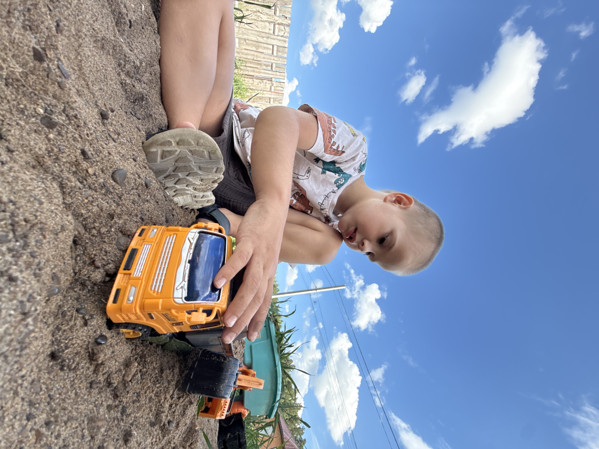 A young child is playing in the sand with a toy truck. The child, wearing a t-shirt with dinosaur print and dark shorts, is kneeling and holding the orange and black toy truck. The child's left foot, clad in a grey sneaker, is visible in the sand. The scene is outdoors with a bright blue sky filled with fluffy white clouds, suggesting a sunny day. Behind the child, a wooden fence is partially visible, and some green foliage and a house roof can be seen in the background, hinting at a backyard or park setting. The perspective is low-angle, looking up towards the sky, making the child and the truck the central focus. The overall mood appears to be one of playful engagement and enjoyment. There is no visible text in the image.