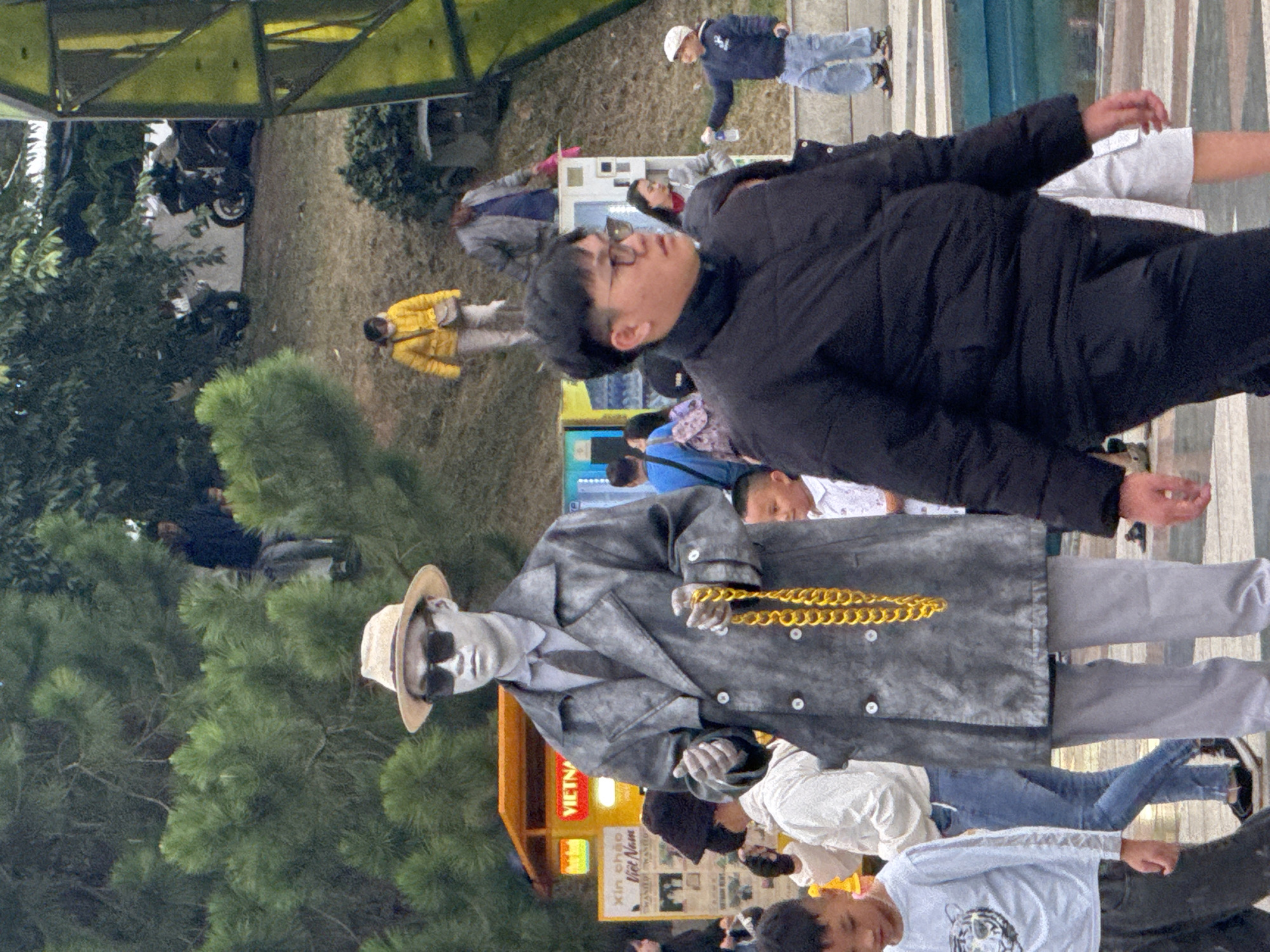 The image shows a street scene in Da Lat, Vietnam. In the foreground, a person dressed as a silver statue stands wearing a grey trench coat, a hat, sunglasses, and a thick gold chain. To the left, a young boy in a light blue long-sleeved shirt walks by. Behind the statue, a woman in a white jacket and jeans is visible. In the background, other people are walking, and a yellow food stall with Vietnamese writing is present. The environment includes trees and some modern structures.