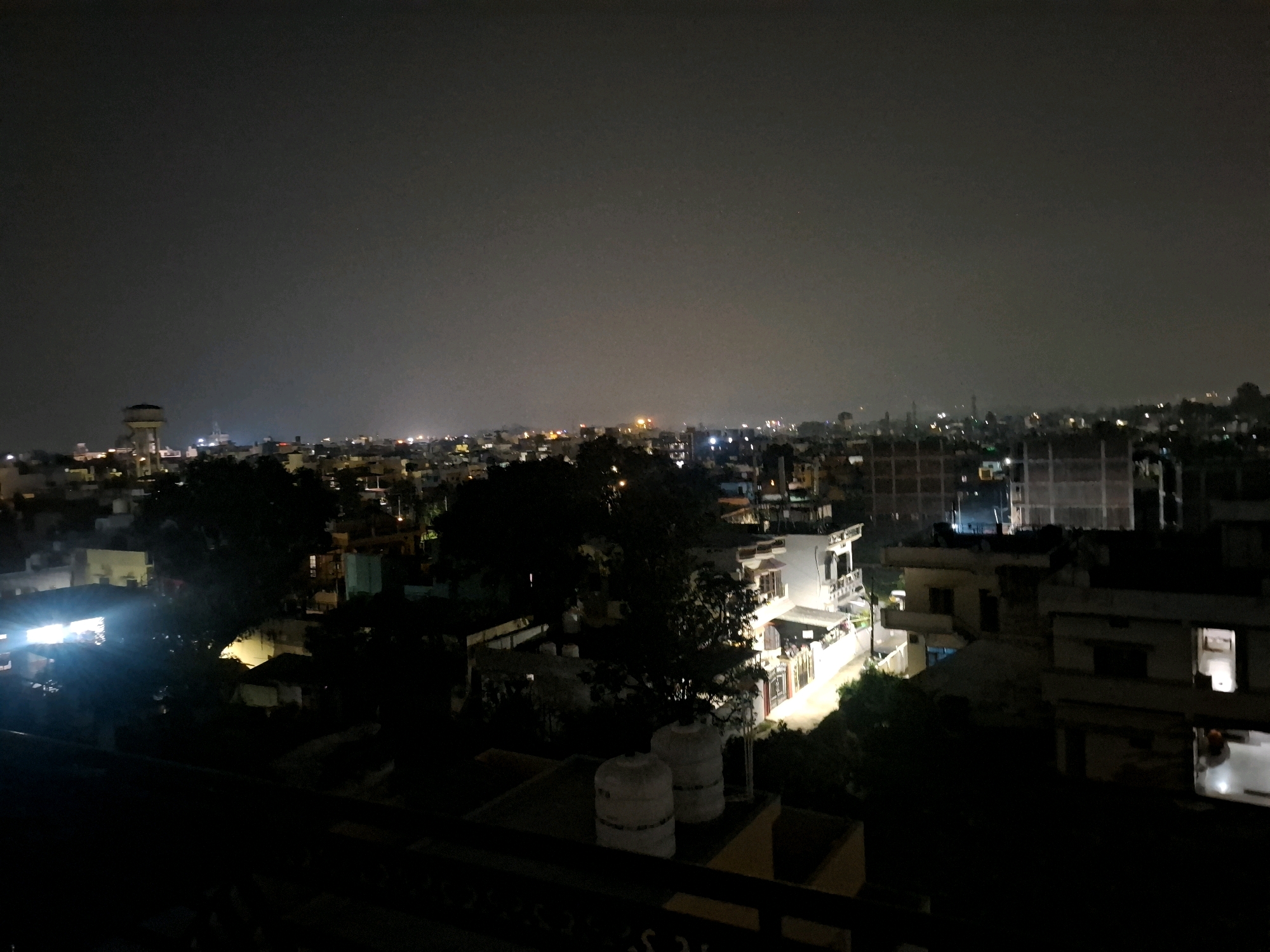 A nighttime aerial view of a densely populated urban area in Dehradun, India shows a cityscape illuminated by artificial lights. Buildings of varying heights and architectural styles are visible, including residential houses, apartment buildings, and what appears to be a water tower in the distance on the left. The sky is dark and overcast, with some faint lights suggesting the presence of distant stars or lights. The foreground is dominated by the rooftops of buildings, including two large white water tanks on one of the closer structures. The overall impression is of a bustling city at night, with a mix of residential and commercial structures.