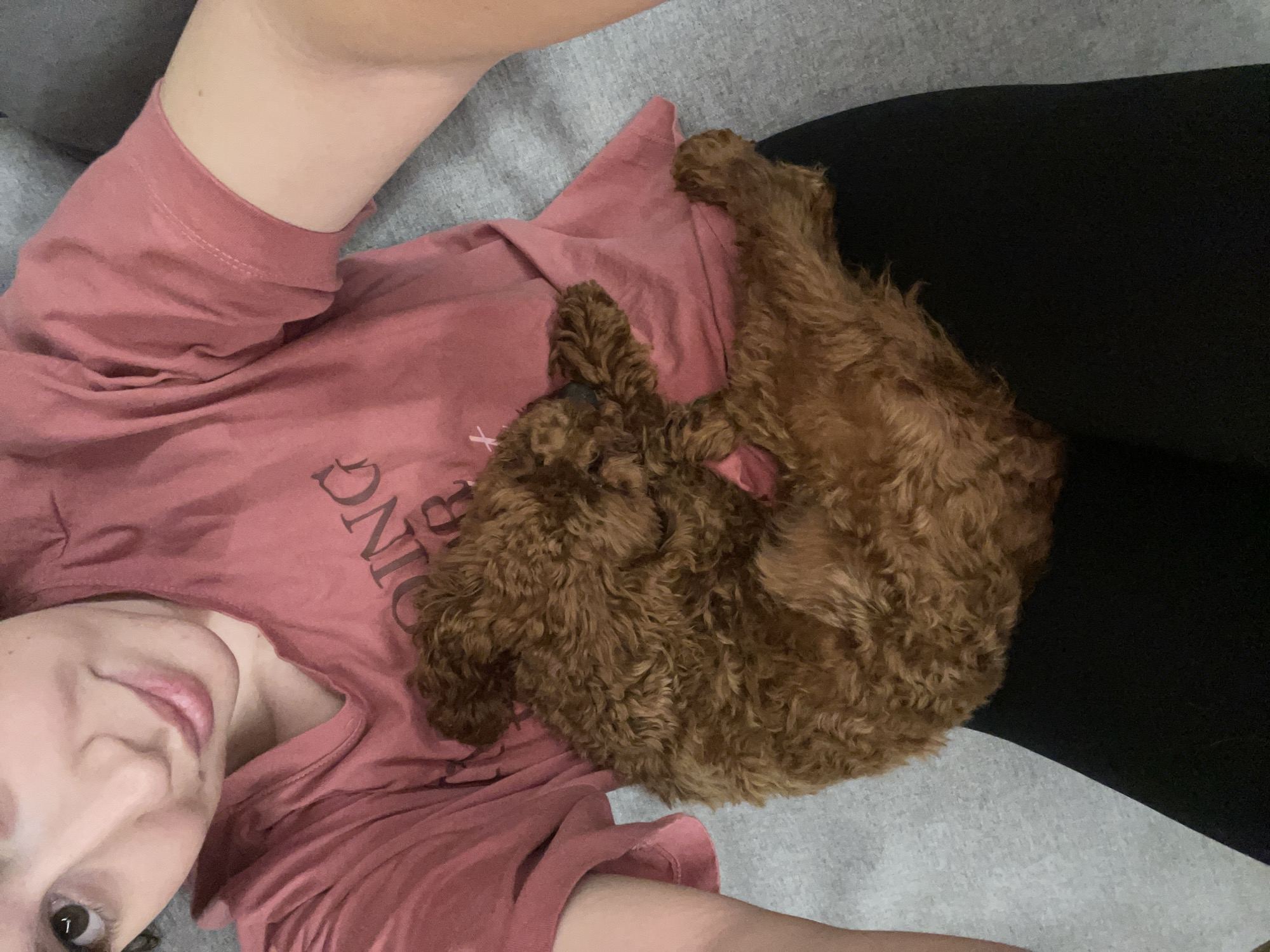 A woman lies on a gray surface, likely a couch or bed, wearing a pink t-shirt and black pants. A small, brown, curly-haired dog is curled up on her chest. The woman is smiling and looking at the camera. The location is unknown and not determinable from the image. There is no evidence to suggest the image was taken in Sofia, Bulgaria.