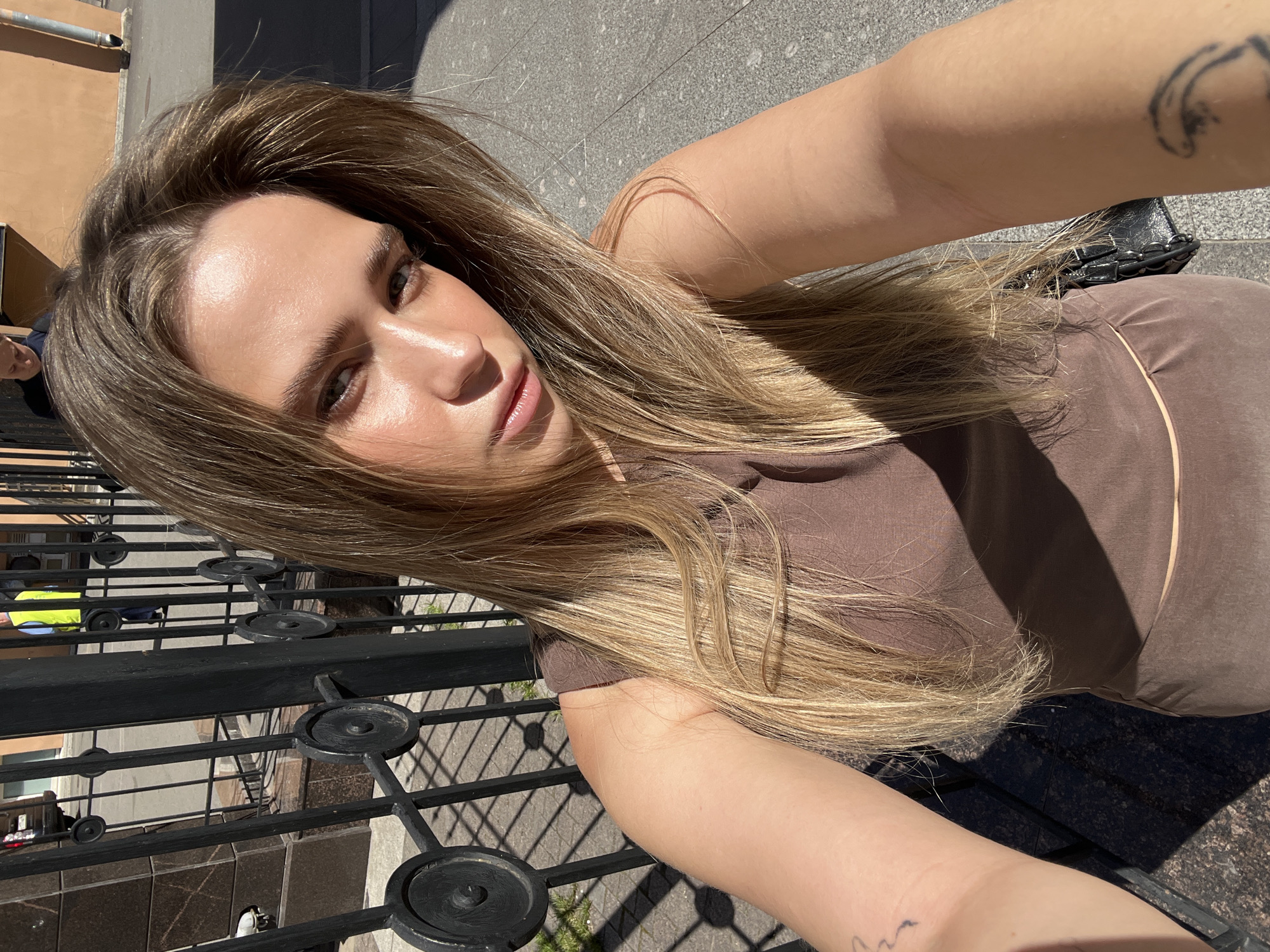 The image is a selfie taken by a young woman with blonde hair, wearing a brown sleeveless top. She is standing outdoors in front of a black metal fence. The background includes a building with a light brown facade and pavement. In the distance, a person wearing a yellow vest and a dark vehicle are visible. There is a shadow cast across the ground, indicating it is daytime. The woman has a tattoo on her left arm and what appears to be the corner of a black purse is visible near her right arm. The setting suggests an urban environment in Saint Petersburg, Russia, possibly near an entrance or public space given the fence.