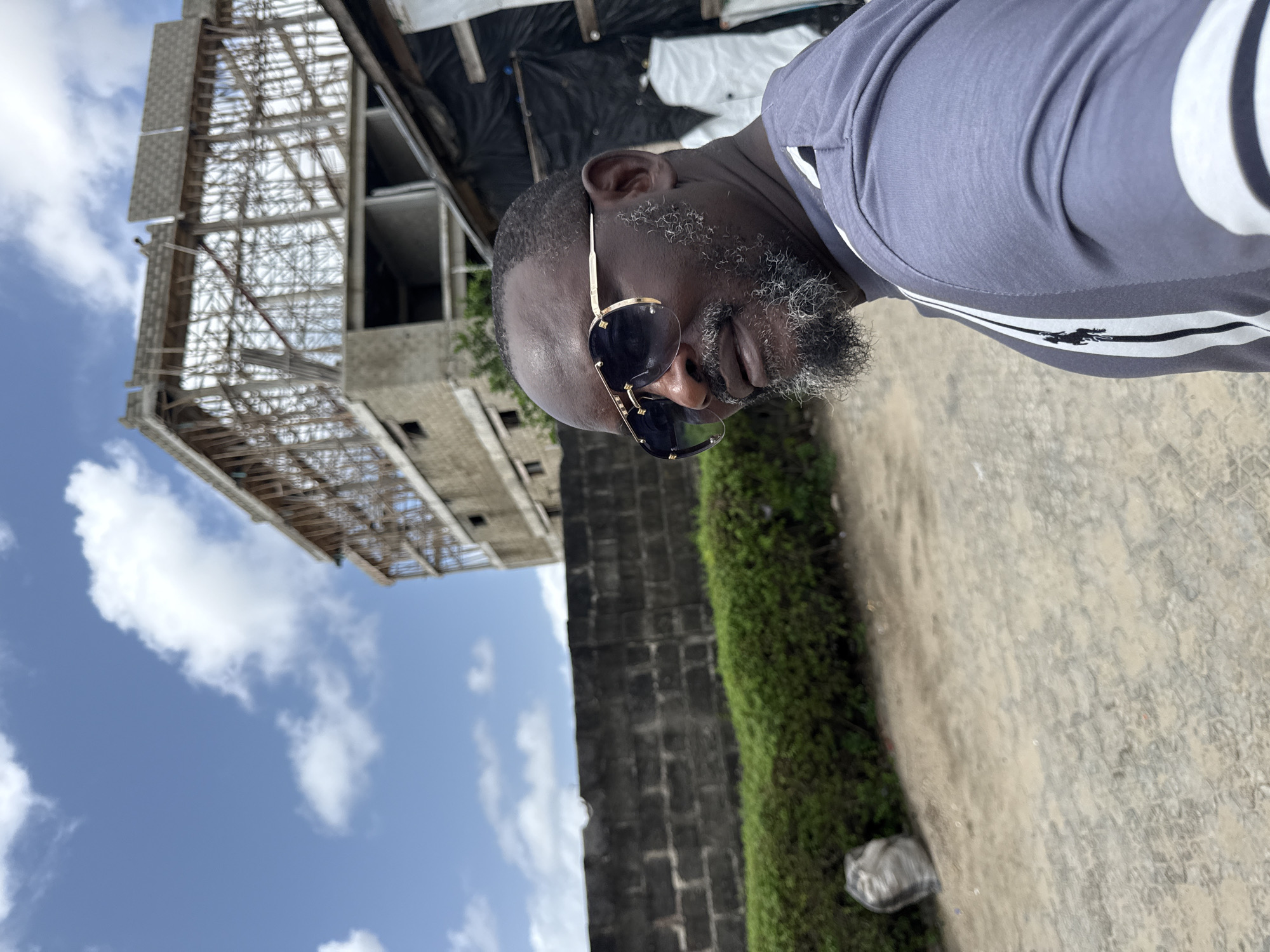 A medium shot of a man in Ado, Nigeria, wearing sunglasses and a gray t-shirt with white stripes, standing on a concrete surface with green vegetation to his right. Behind him is a tall brick wall, a building under construction, and a partially cloudy blue sky.
