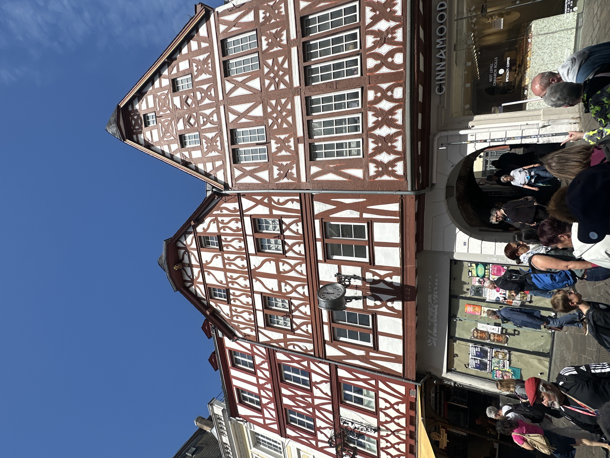 The image depicts a bustling street scene in an unknown city under a clear, bright blue sky with sparse wispy clouds visible on the right. The prominent architectural features are three multi-story half-timbered buildings dominating the midground, characterized by their reddish-brown structural timbers and white or off-white infill panels. The buildings display varied intricate timber patterns, including geometric, wavy, and crisscross designs. They are fitted with numerous rectangular windows featuring white frames, often divided into multiple panes. The roofs are steep and dark, with the rightmost building exhibiting a tall, ornate gable adorned with detailed timbering and a decorative finial at its apex.
A white, circular clock with black Roman numerals and hands is mounted on a decorative bracket on the facade of the central building, casting a shadow. Below it, the faded numbers "60Z" are visible. A dark, vertical drainpipe runs down the face of the leftmost half-timbered building.
At street level, the ground floors host commercial establishments. On the left, a yellow awning extends over a storefront, while the adjacent shop window displays various posters for events such as DOTA, music, and comedy. On the right, a large glass window reveals a shop named "CINNAMOOD," displaying cinnamon rolls and listing "NEXT LEVEL CINNAMON ROLLS" and "SPECIALTY COFFEE," along with opening hours. An arched passage extends beneath the building between the central and rightmost structures. A dark utility pole covered in stickers stands near the archway.
The paved street in the foreground and midground is populated by numerous pedestrians, primarily adults of diverse ages, including elderly individuals and at least one child. They are casually dressed in everyday attire and are engaged in walking, conversing, and observing the surroundings. Notable individuals include a man in the foreground left with a white beard and a red beret, wearing a black Adidas track jacket with a lanyard. Near the "CINNAMOOD" shop, a person in a bright green and black patterned jacket is visible making a peace sign with their hand. Many individuals carry backpacks or handbags.