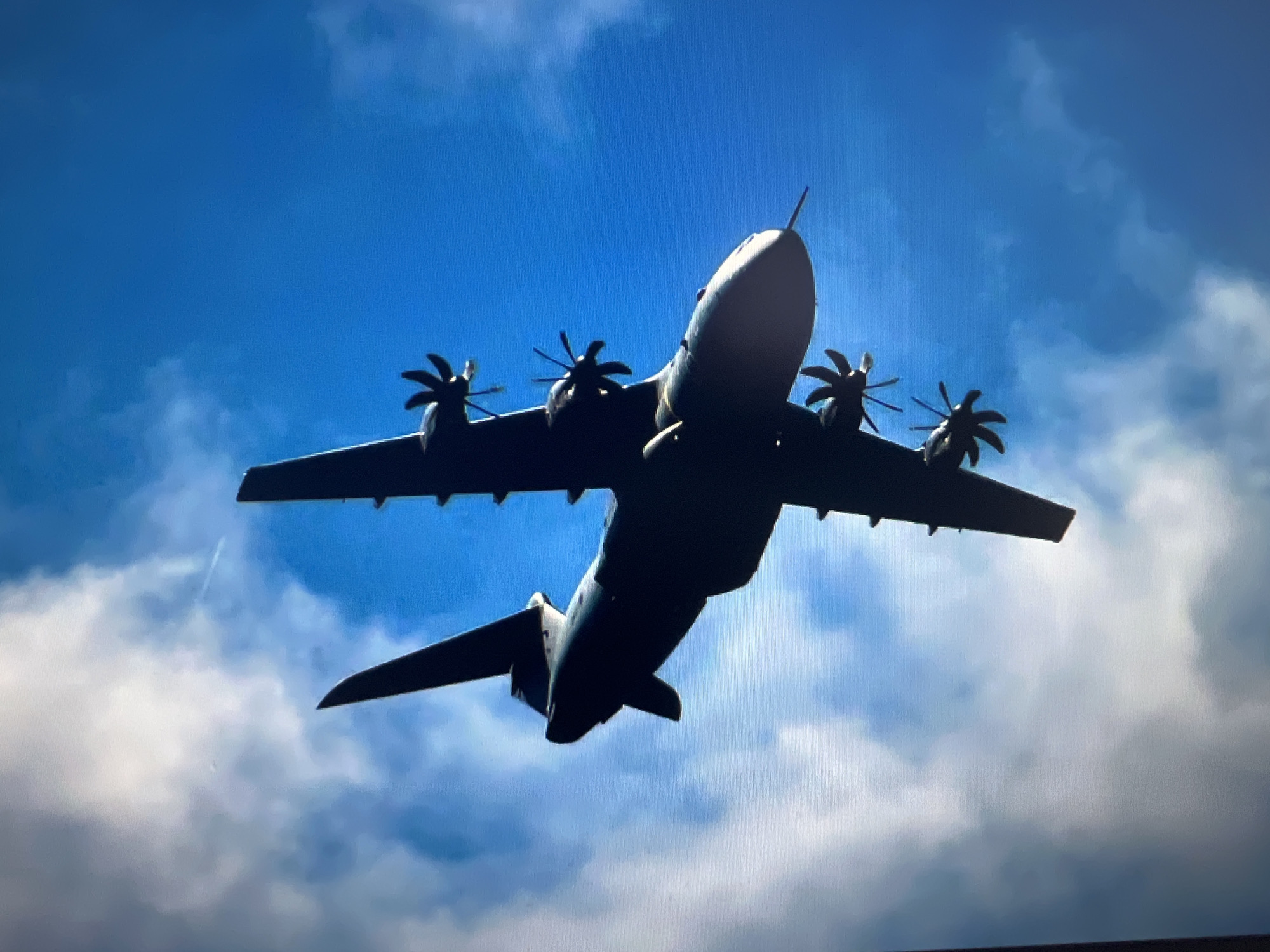 An upward-angle photograph captures a large, four-engine turboprop aircraft in flight, silhouetted against a bright blue sky with scattered white clouds. The aircraft, resembling an Airbus A400M Atlas due to its distinctive shape and eight-bladed propellers on each engine, is seen from directly below, its underside dark against the brighter sky. A prominent probe extends from the nose of the aircraft. The propellers are visible, suggesting they are in motion, though motion blur is not distinct. The wings extend widely, and the tail section is also visible, maintaining the dark silhouette. The sky transitions from a deeper blue at the top to a lighter hue where wispy white clouds are present, particularly towards the bottom and right side of the frame. There are no people visible, and no discernible text on the aircraft. The scene depicts a clear, bright daylight sky, possibly over Varna, Bulgaria, given the provided context.