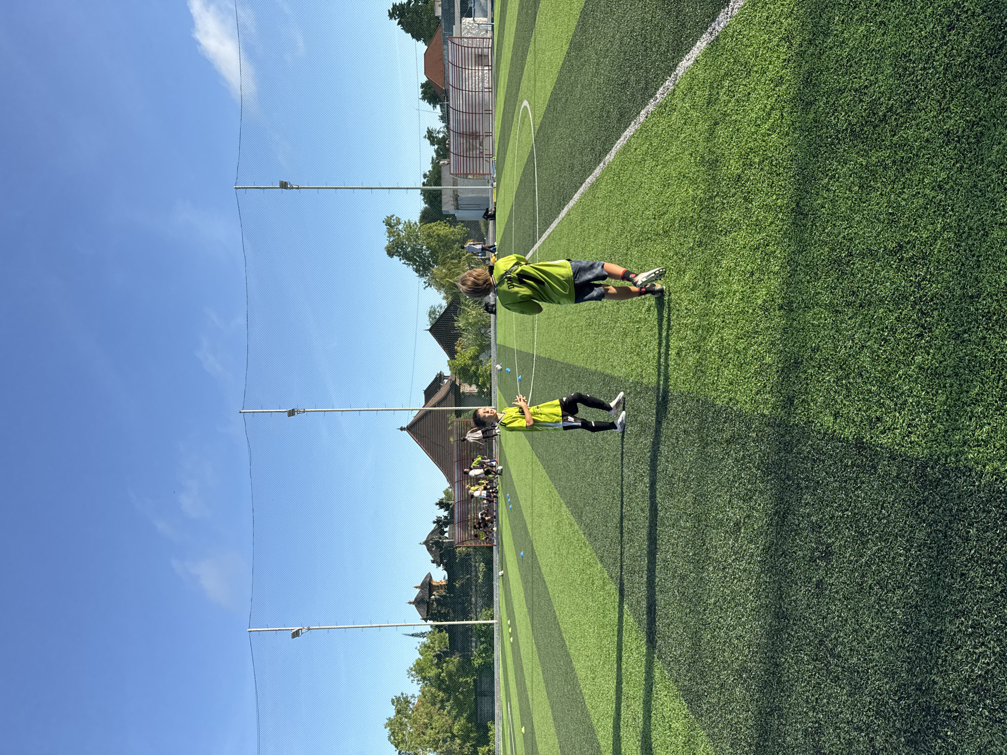 Two children wearing athletic clothing are positioned on a striped green athletic field. The foreground child wears a light green top with black bottoms. The background child also wears a light green top, with dark gray bottoms. Both are wearing cleats. A green mesh net on poles runs along the left edge of the field, under a clear blue sky. Beyond the field are buildings and vegetation, defining the perimeter of the athletic space. Given the buildings and vegetation, this is likely located in Kuta Selatan, Indonesia.