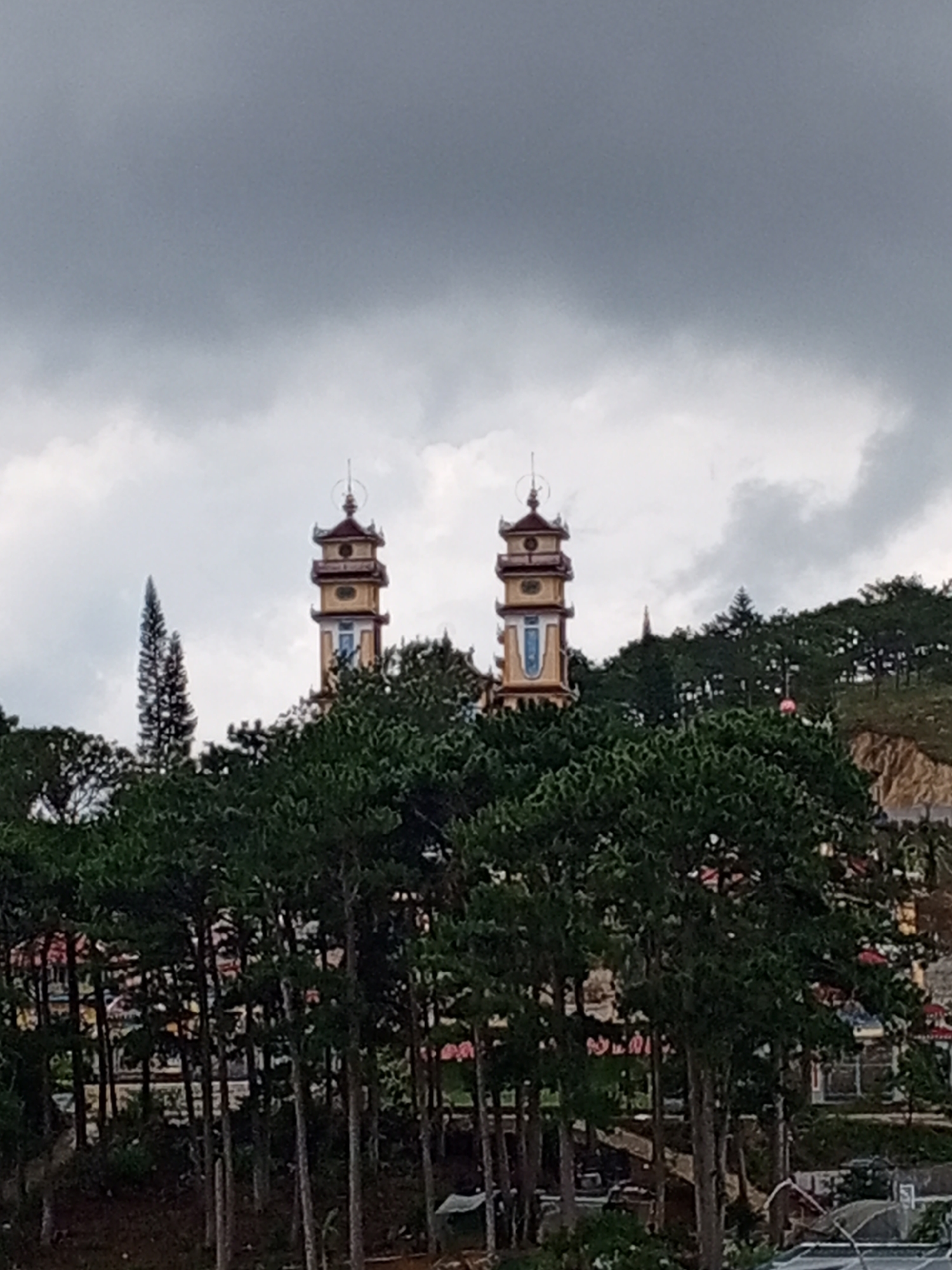 The image depicts a view of two pagodas rising above a dense cluster of pine trees. The sky is overcast with dark, heavy clouds. The pagodas, one slightly taller than the other, are ornately decorated with yellow and red accents and feature multiple tiered roofs. They are situated on a hillside covered in greenery. In the foreground, the dark green foliage of pine trees dominates the lower half of the frame, obscuring a portion of the landscape and any potential structures below. The overall scene suggests a temple or religious site nestled within a natural, possibly mountainous, environment. The location is identified as Xuan Thanh, Vietnam.