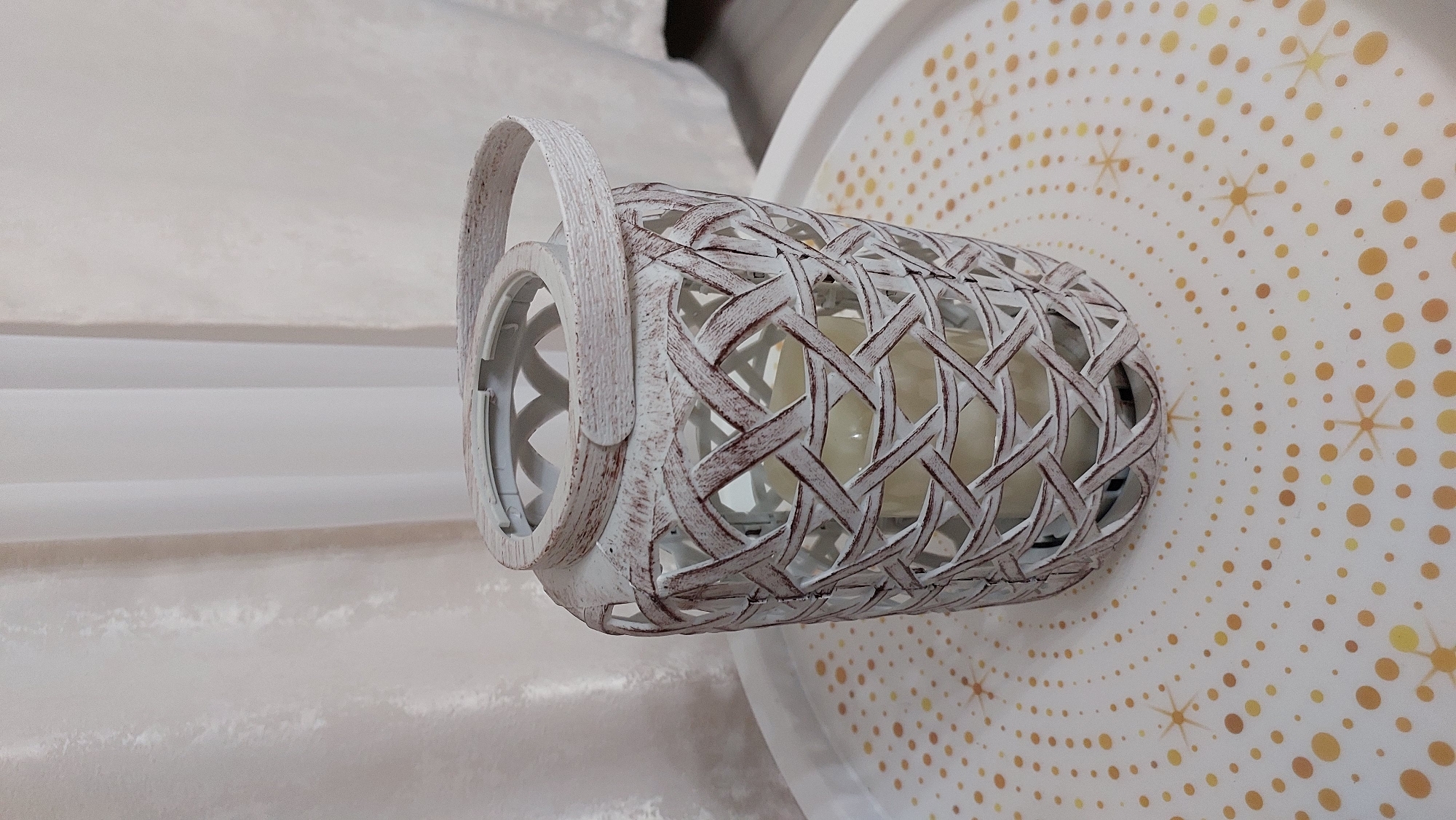 An antique white lattice lantern with a single candle inside sits on a white tray decorated with gold polka dots and starbursts. The lantern has a woven appearance and a distressed finish, with hints of dark wood showing through the white paint. A curved handle is attached to the top rim of the lantern. The tray is positioned on a surface in front of a light-colored, textured curtain. The image is a close-up, focused on the lantern and tray, with the background softly blurred. The scene is indoors and appears to be a still-life arrangement. There is no indication of Novocheboksarsk, Russia.