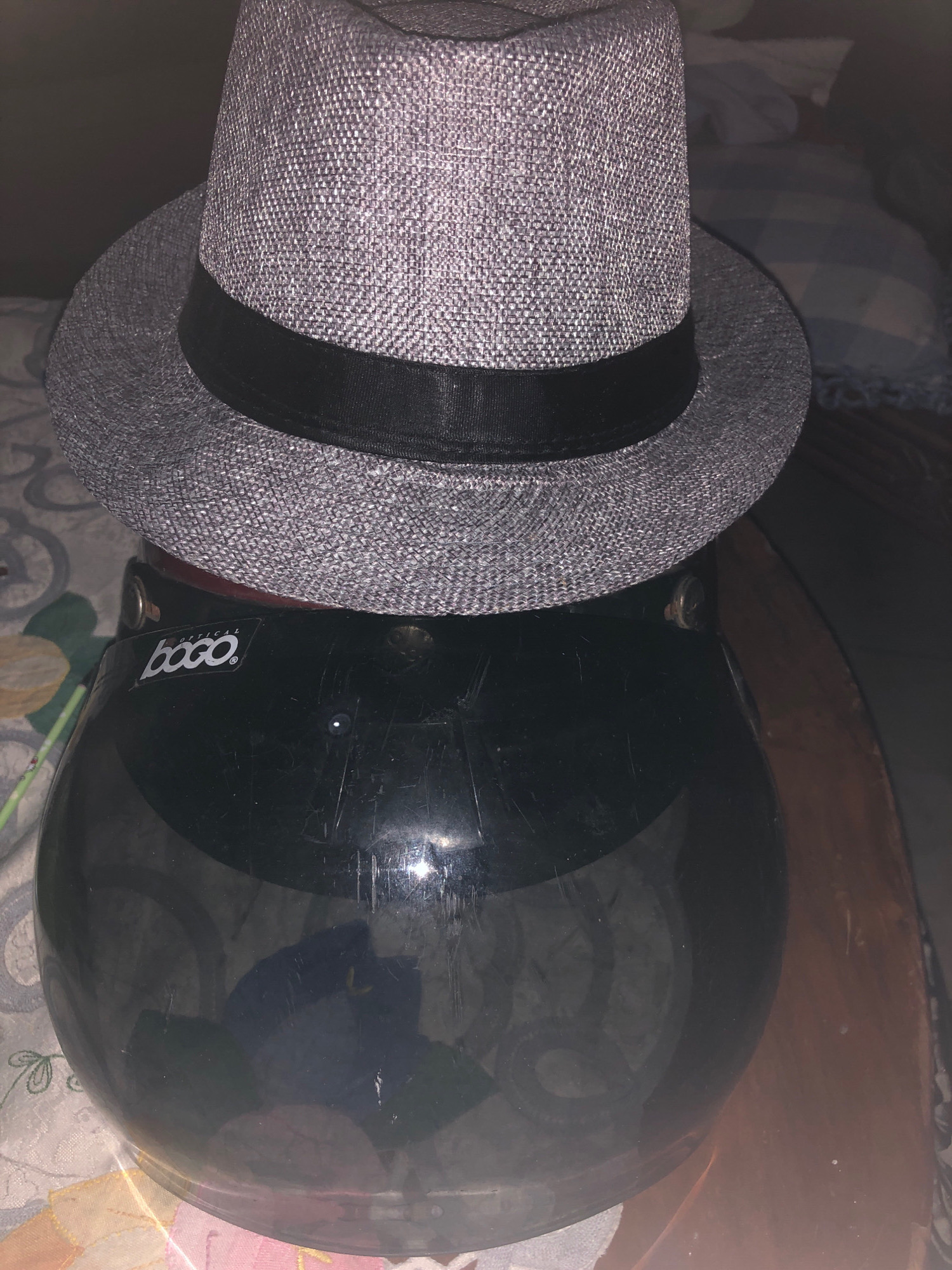 A grey fedora hat sits atop a black motorcycle helmet. The hat has a black band around its base. The helmet has a reflective visor, and a sticker with the word "bogo" is visible on the side. The background appears to be a bed with patterned sheets, suggesting an indoor setting. The image is likely taken in Lhokseumawe, Indonesia, given the context provided. There are no people present in the image.