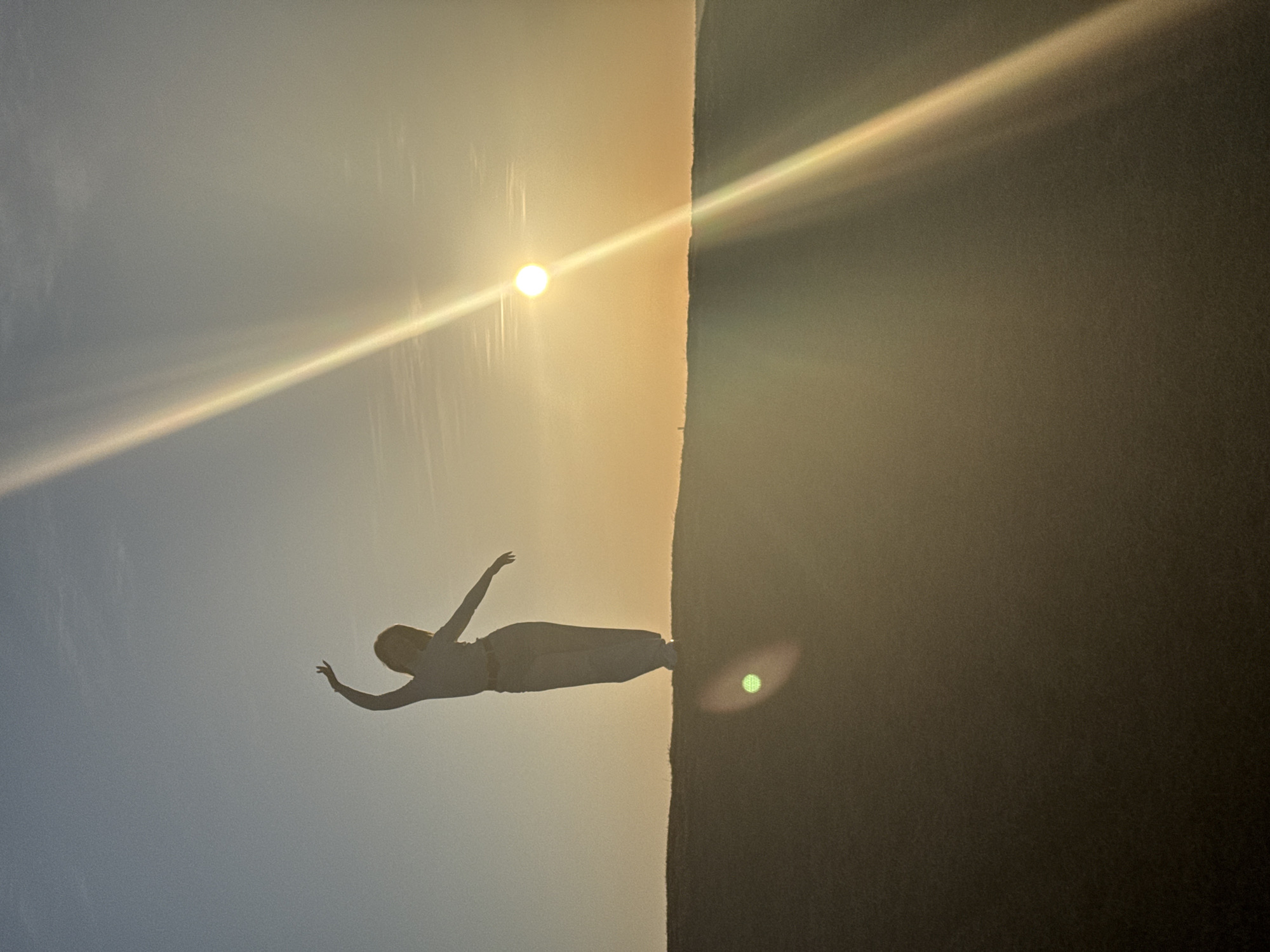 A long shot shows a person silhouetted against a sky with a bright light source (likely the sun) and lens flares. The sky transitions from a lighter hue in the upper left to a darker hue on the right, with a visible edge separating the two. The person is positioned horizontally with arms outstretched, possibly performing a dance-like or acrobatic movement. The location in the city of Ольховка, Russia is not discernible from the image.