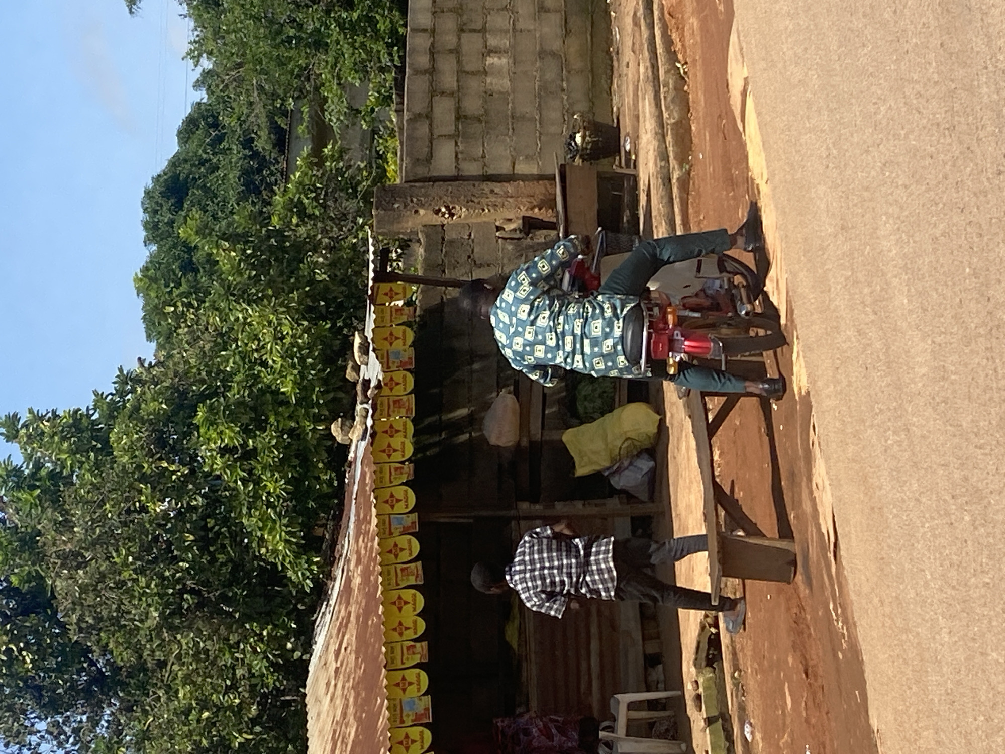 In Gura Top, Nigeria, a young boy stands facing away from the camera, dressed in a black and white checkered shirt and dark pants. He is positioned in front of a roadside stall with a corrugated metal roof and yellow banners. To his right, a man sits on a red motorcycle, also facing away from the camera. He is wearing a green and white patterned long-sleeved shirt and dark pants. The motorcycle is parked on a dirt and gravel area next to a paved road. Behind the stall and the motorcycle, a concrete wall is visible. Lush green trees and foliage form a backdrop, suggesting a rural or semi-urban setting. The sky above is clear and blue.