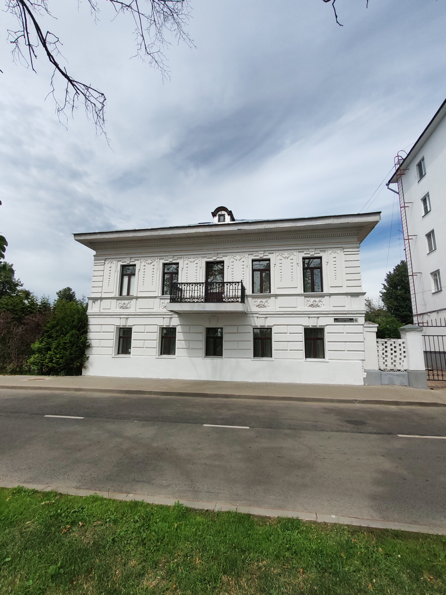 A three-story white building with a dark roof and a balcony is visible on a cloudy day in Rusanovo, Russia. The building has ornate white detailing around the windows and along the roofline. A dark wrought-iron balcony with a decorative railing extends from the second story. The building is situated next to a gray asphalt road with white lane markings, bordered by a strip of green grass. A taller white building with a metal fire escape is visible to the right of the main structure. A decorative white fence with a black gate stands in front of the building on the right side. Trees are visible behind the building and on the left side of the image. The sky is overcast with some wispy clouds.