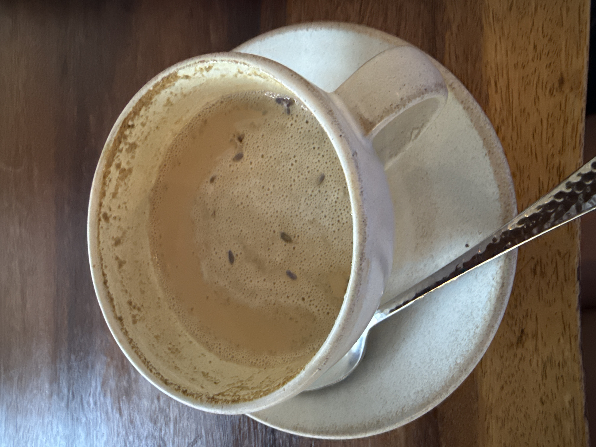 A top-down, close-up shot of a ceramic coffee cup resting on a matching saucer with a spoon placed on the side, set against a background of wooden surfaces. The coffee cup is nearly empty, containing only a small amount of foamy, light brown liquid. There are a few small, dark particles floating in the liquid. Residue is visible along the inside of the cup, indicating where the liquid level was before. The saucer is off-white or beige, with a slightly darker rim. The spoon is metal, with a patterned handle. The setting is likely a table, with portions of the table's wooden surface visible. The location in Canggu, Indonesia cannot be verified from the image alone.
