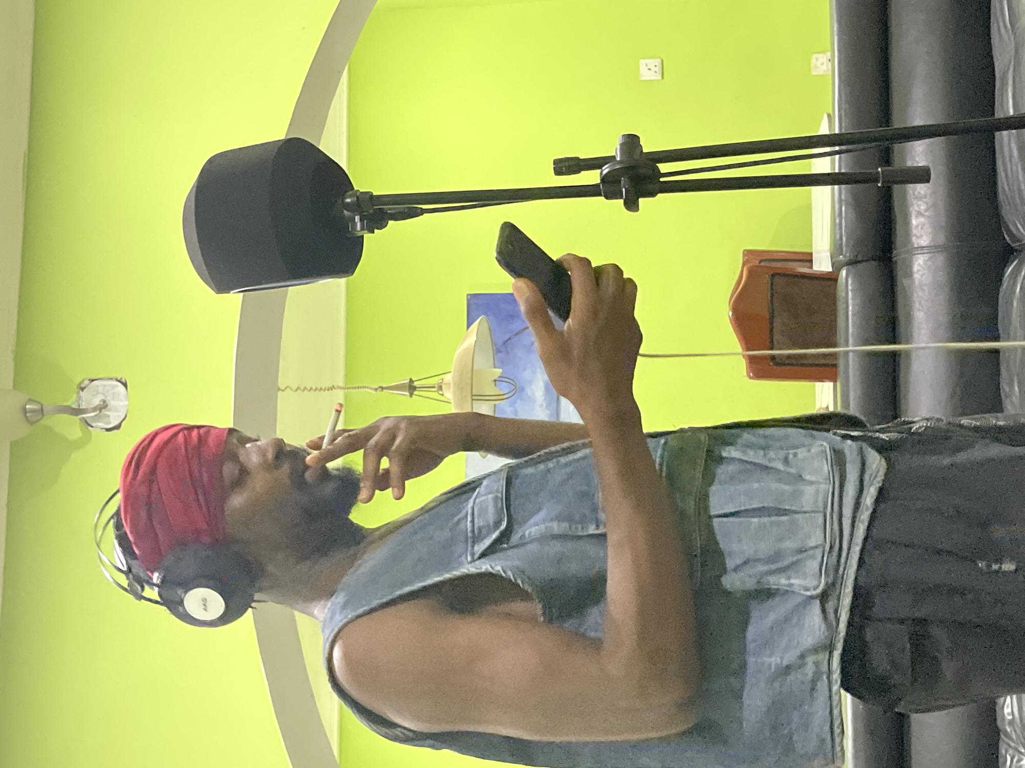 A medium shot depicts a man inside a soundproof recording studio in Abuja, Nigeria. The man, wearing a red headscarf and black headphones, is holding a mobile phone in his right hand and smoking a cigarette held in his left. He has a beard and is wearing a denim vest over a black shirt. A black microphone with a cylindrical pop filter on a stand is positioned in front of him. The walls are a bright lime green. Behind the man is a stack of black foam soundproofing panels. Other visible objects include a light fixture, a small cabinet, and electrical outlets.