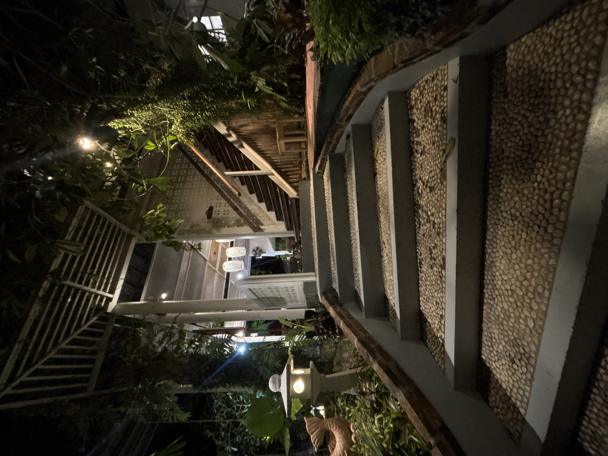 The image shows an outdoor staircase leading up to a building at night. The steps are made of concrete with a surface of small, light-colored pebbles. The staircase is flanked by brick retaining walls and lush green vegetation. A Japanese-style stone lantern with a lit interior stands on the left side of the lower steps. The building appears to have an open-air structure with a slatted roof and white pillars. A wooden staircase with a light-colored handrail is visible to the right of the main staircase, leading up to a platform. Hanging lanterns are visible under the slatted roof. The scene suggests a tropical setting, possibly a restaurant or a resort in Chalong, Thailand, given the architectural style and abundant greenery. There are no people visible in the image.