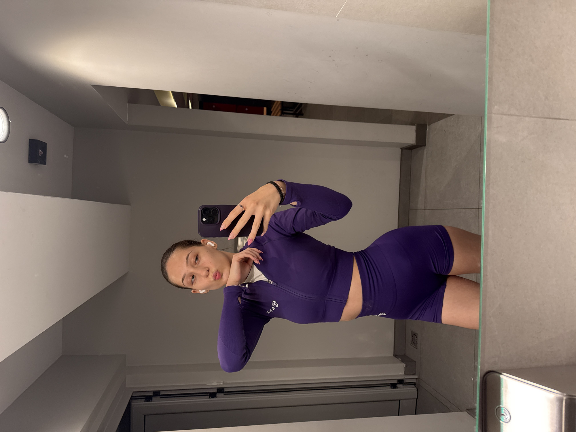 A young woman in a purple athletic set is taking a mirror selfie in a gym locker room. She is wearing a cropped zip-up jacket and matching high-waisted shorts. Her hair is pulled back into a ponytail, and she has earbuds in. She is holding a purple iPhone in her right hand, which she is using to take the picture. The locker room has grey tiled floors and walls, with a metal sink visible in the foreground of the mirror. In the background, there are lockers and a doorway leading to another area of the gym.